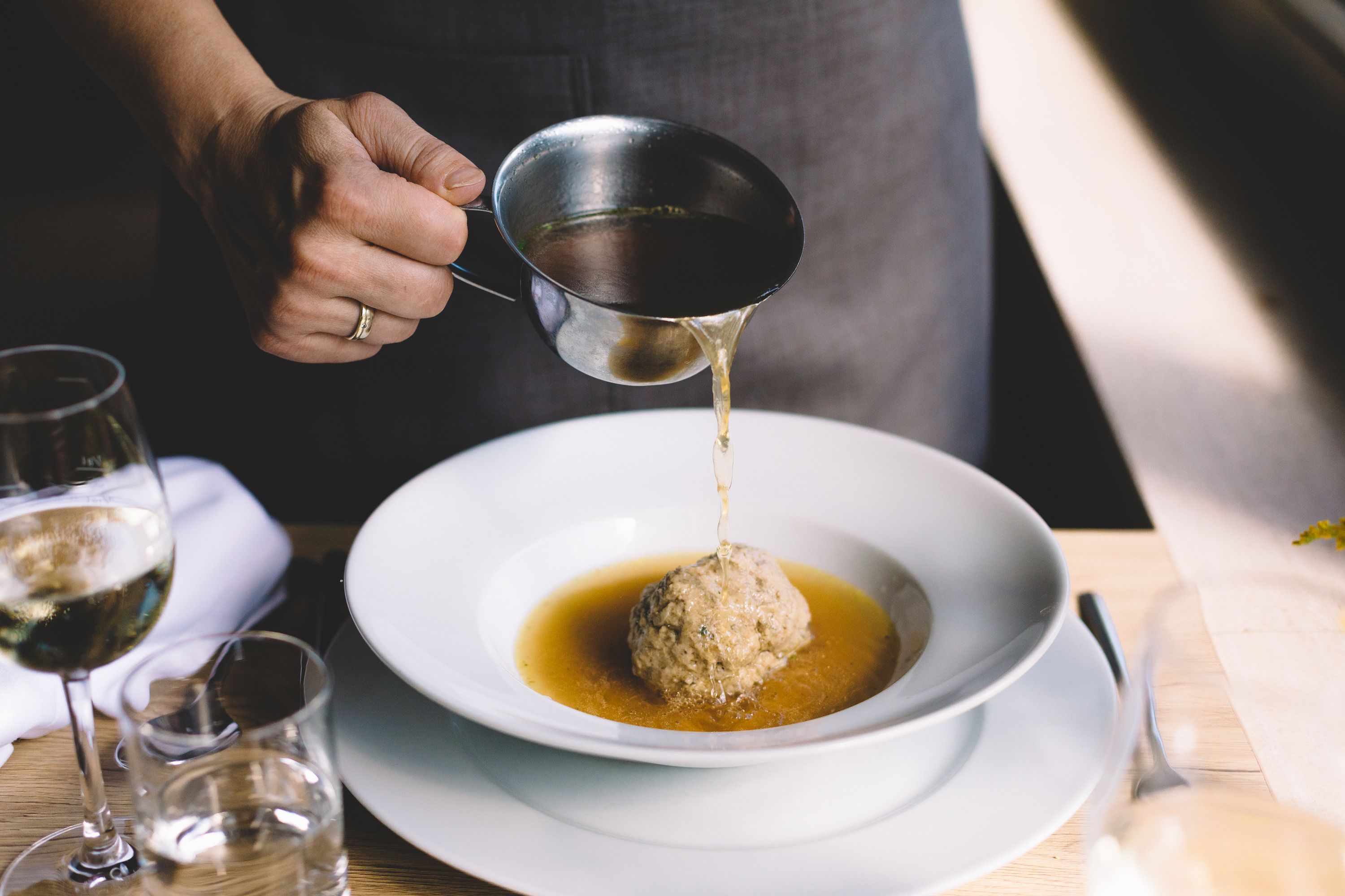 A person pours broth over a liver dumpling in a white plate.