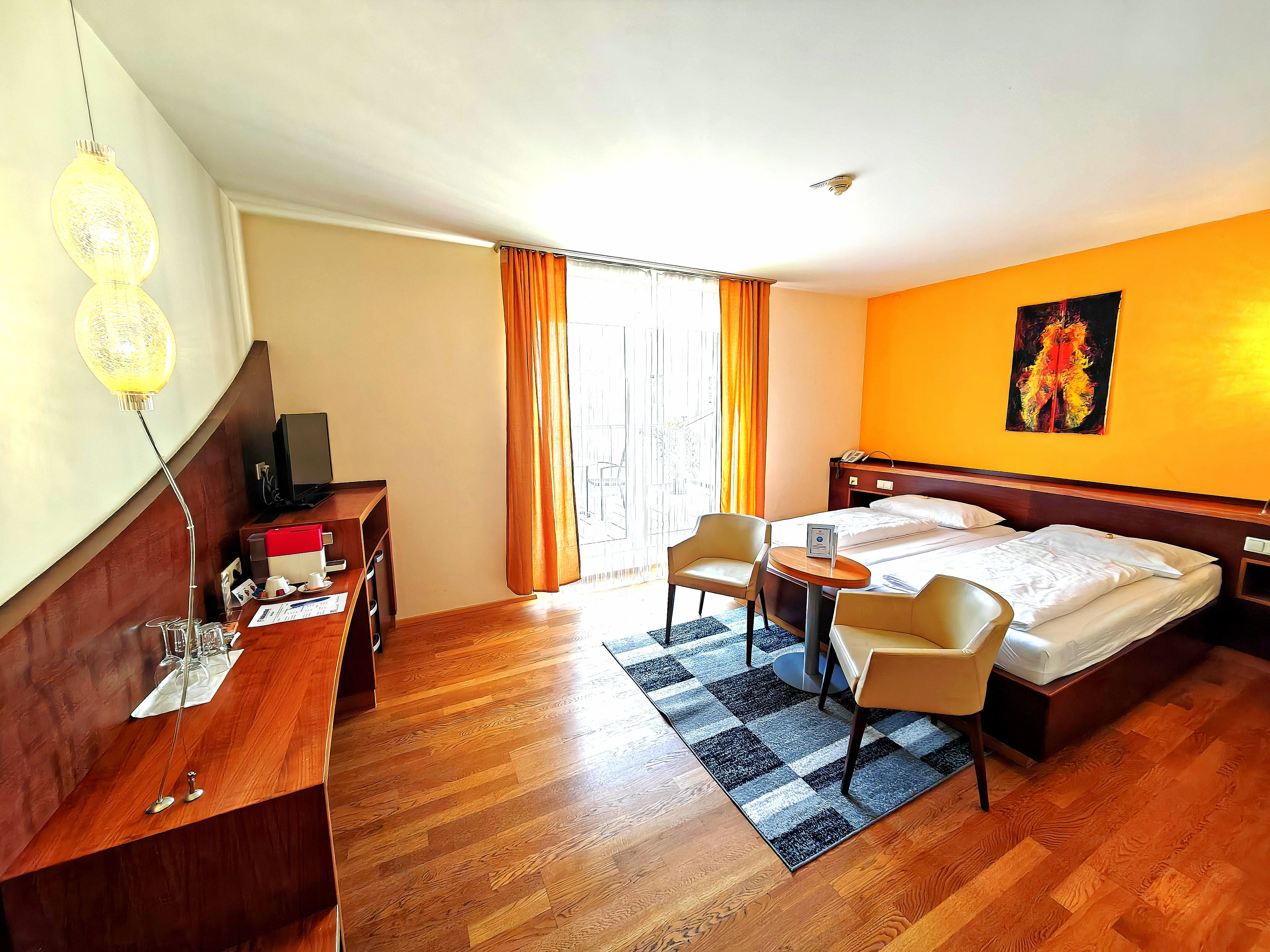 A modern hotel room with a double bed, two armchairs, a small table, and a desk with a TV. The walls are painted in warm colors.