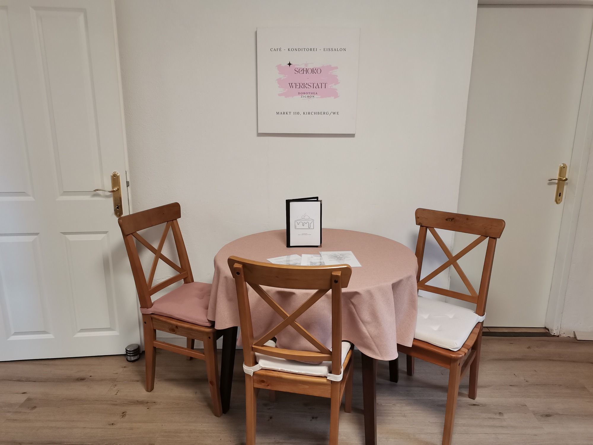 A table with a pink tablecloth and three wooden chairs in a café, with a sign on the wall.