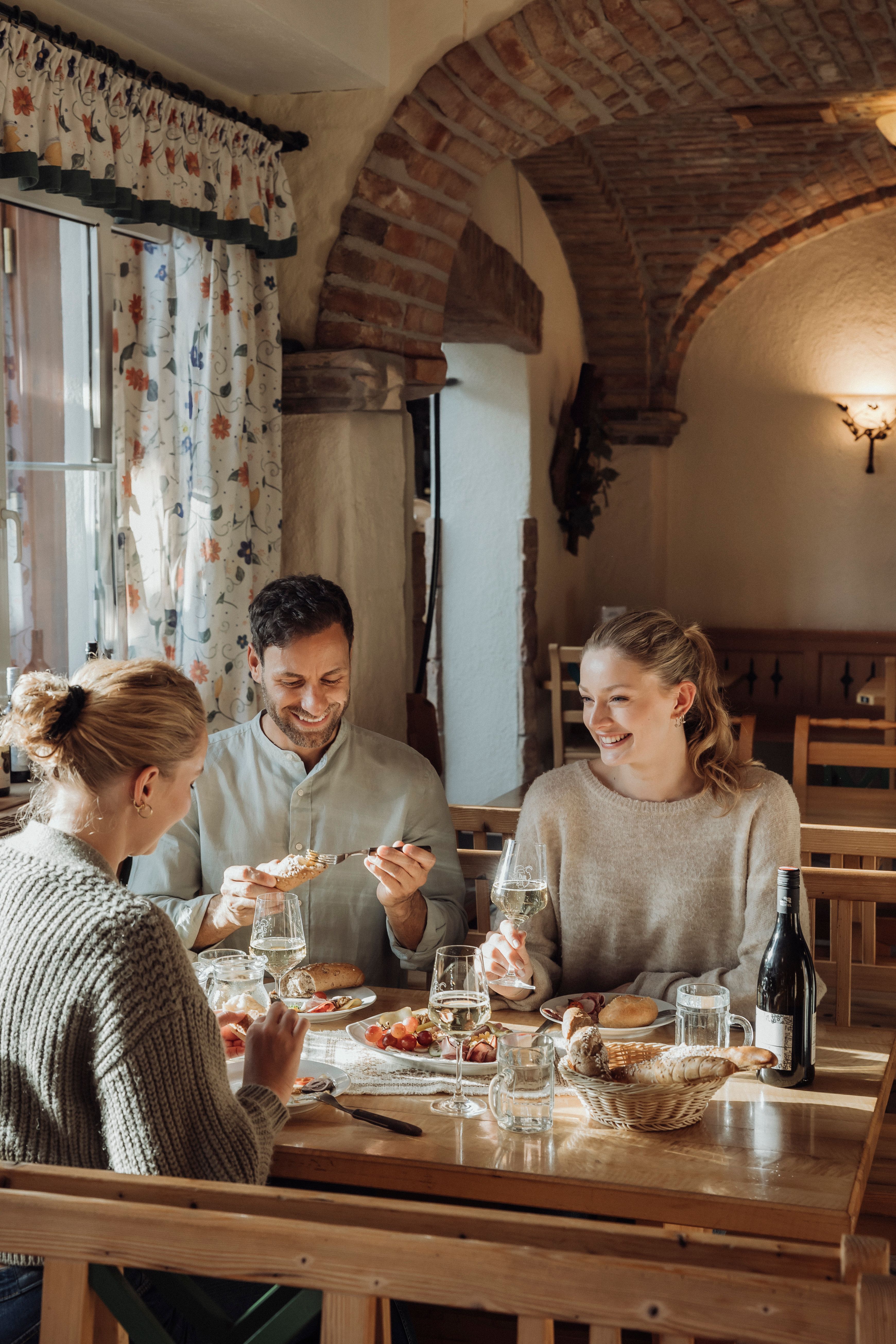Three people sit at a table in a cozy restaurant and enjoy wine and food.