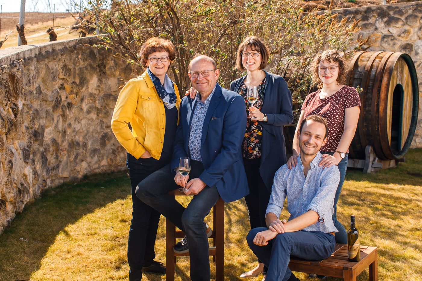 A smiling family poses outdoors in front of a stone wall with a wine barrel in the background.