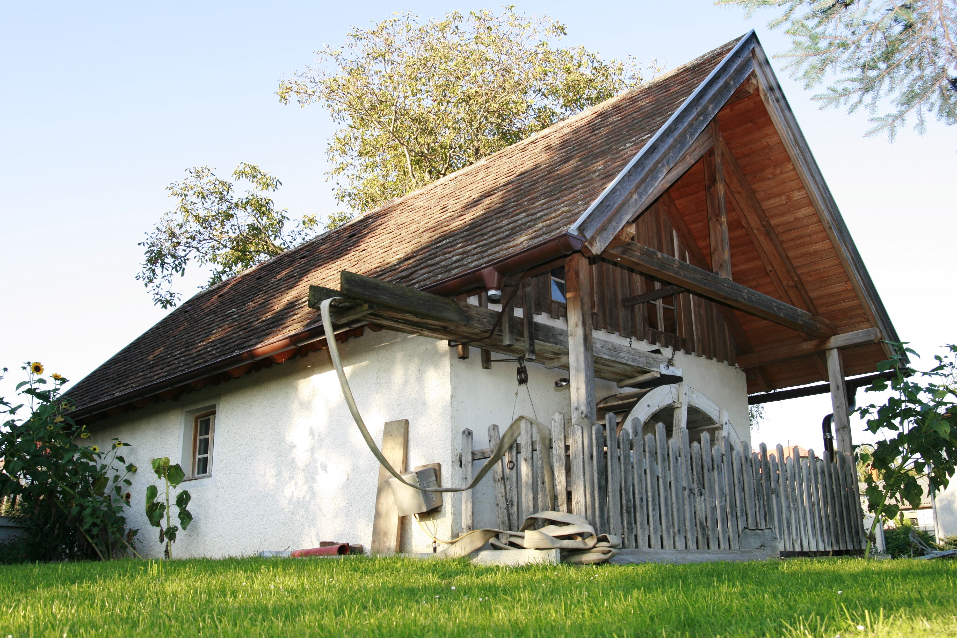 Traditional building with wooden roof and fence, surrounded by green meadow and trees.
