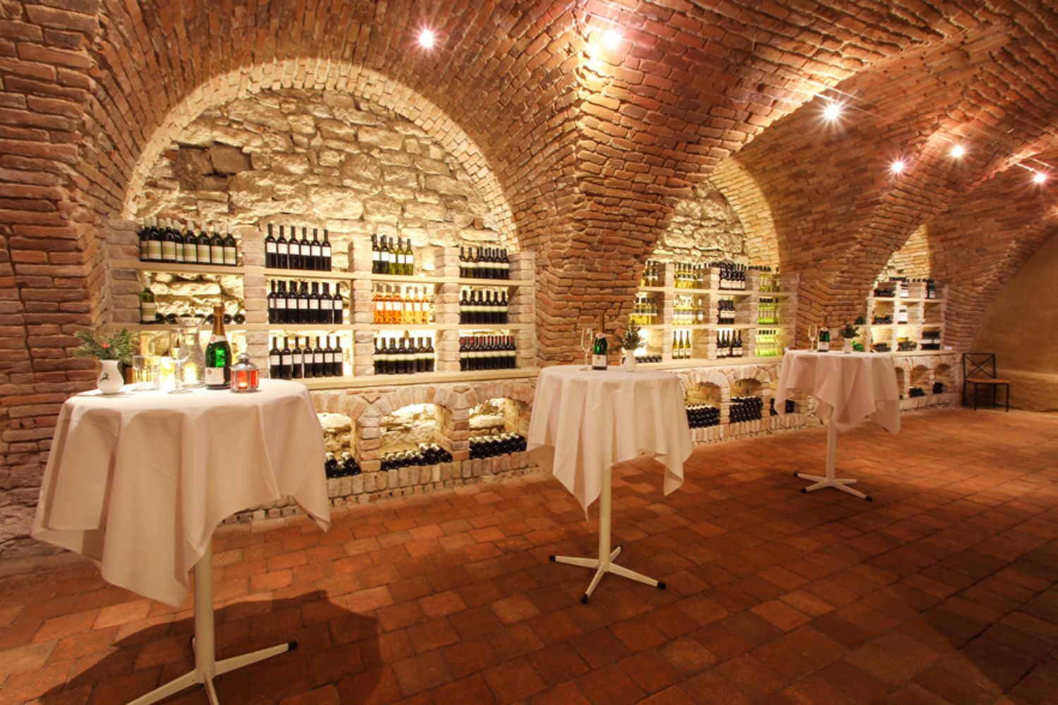 A wine cellar with brick walls, shelves full of wine bottles and bar tables with white tablecloths.