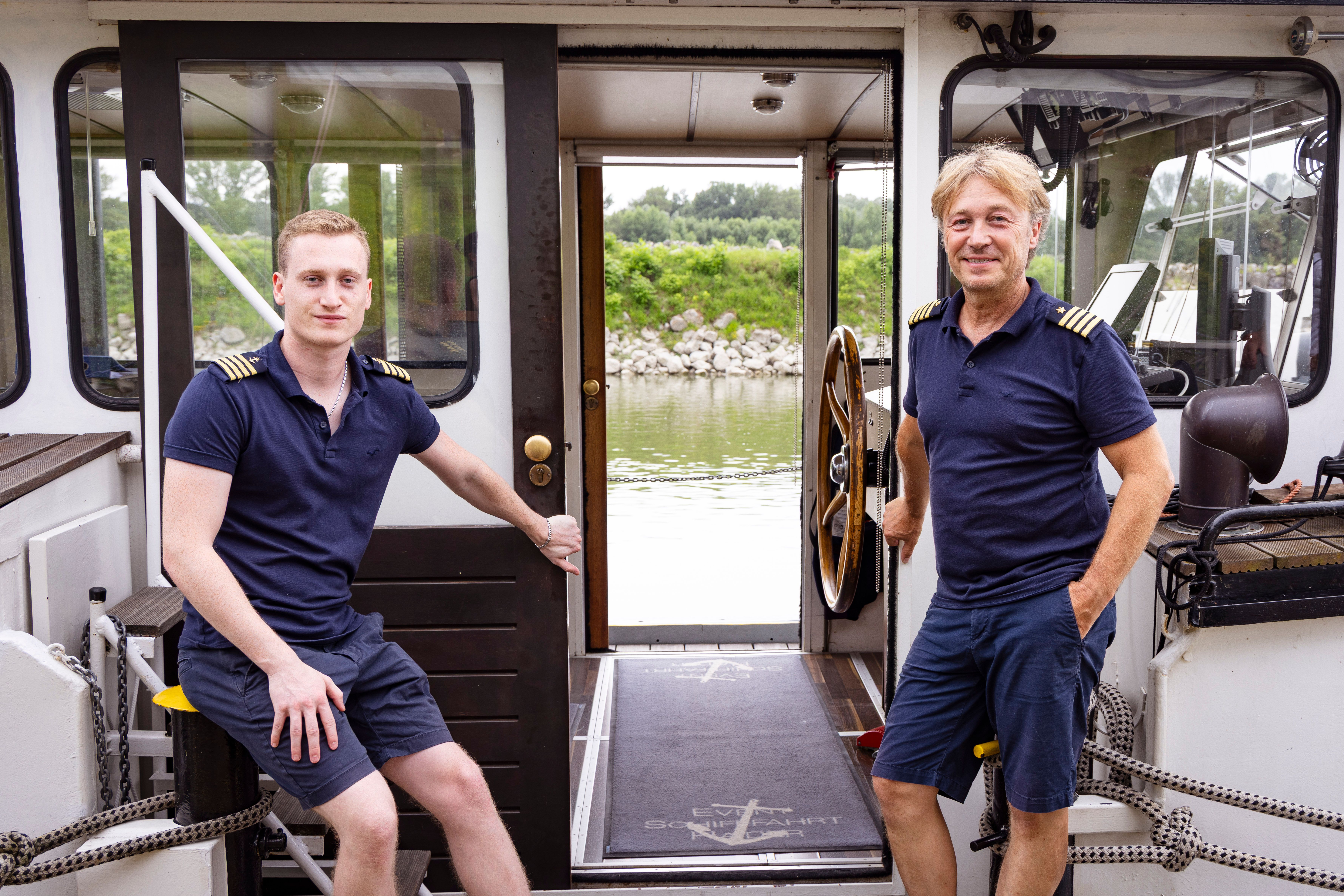 Two men in naval uniform on a ship, with a view of a river and green shore landscape.