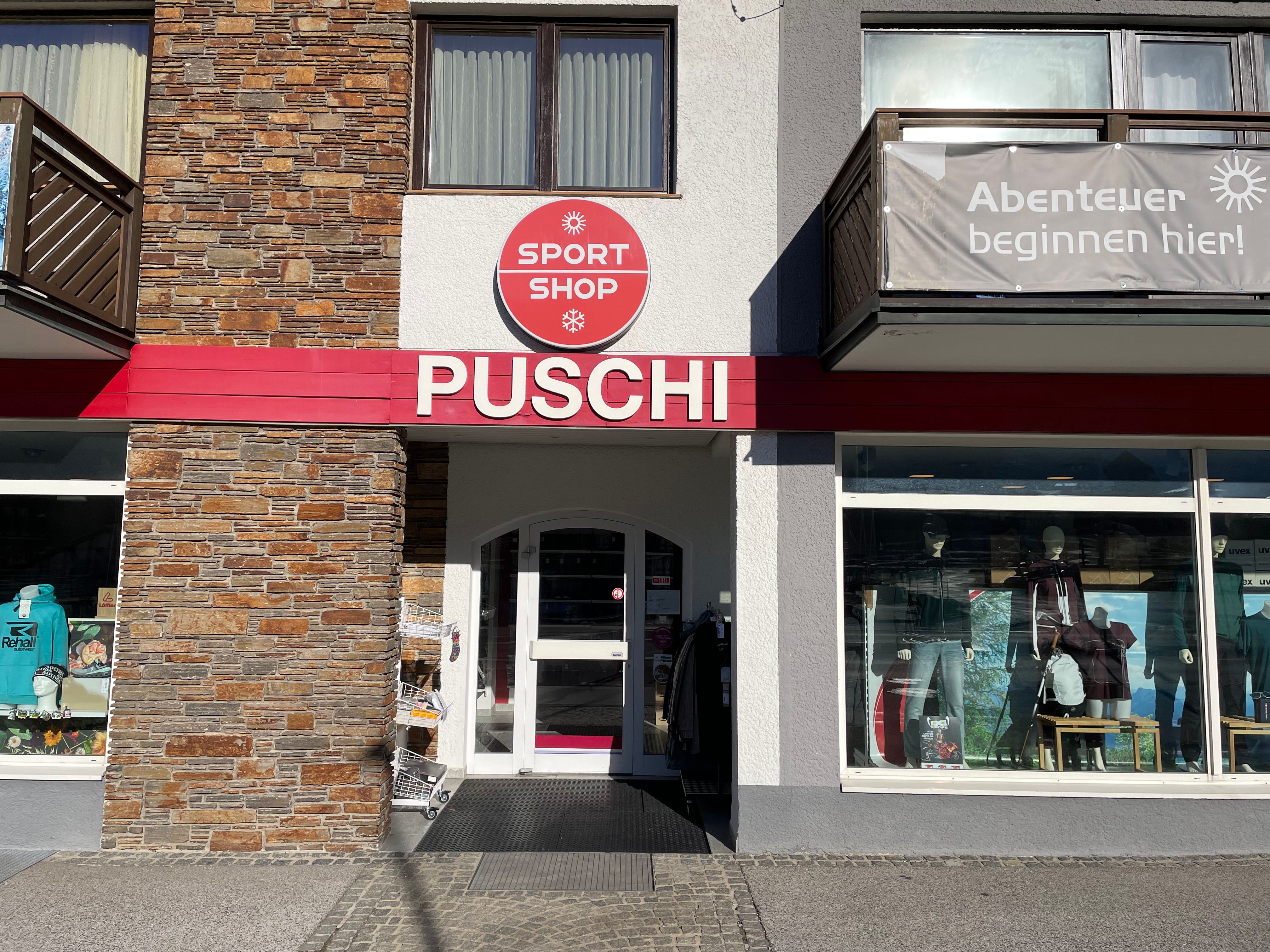 Entrance area of a sports store with the inscription 'PUSCHI' and a sign 'SPORT SHOP'. Shop window with sportswear and equipment.