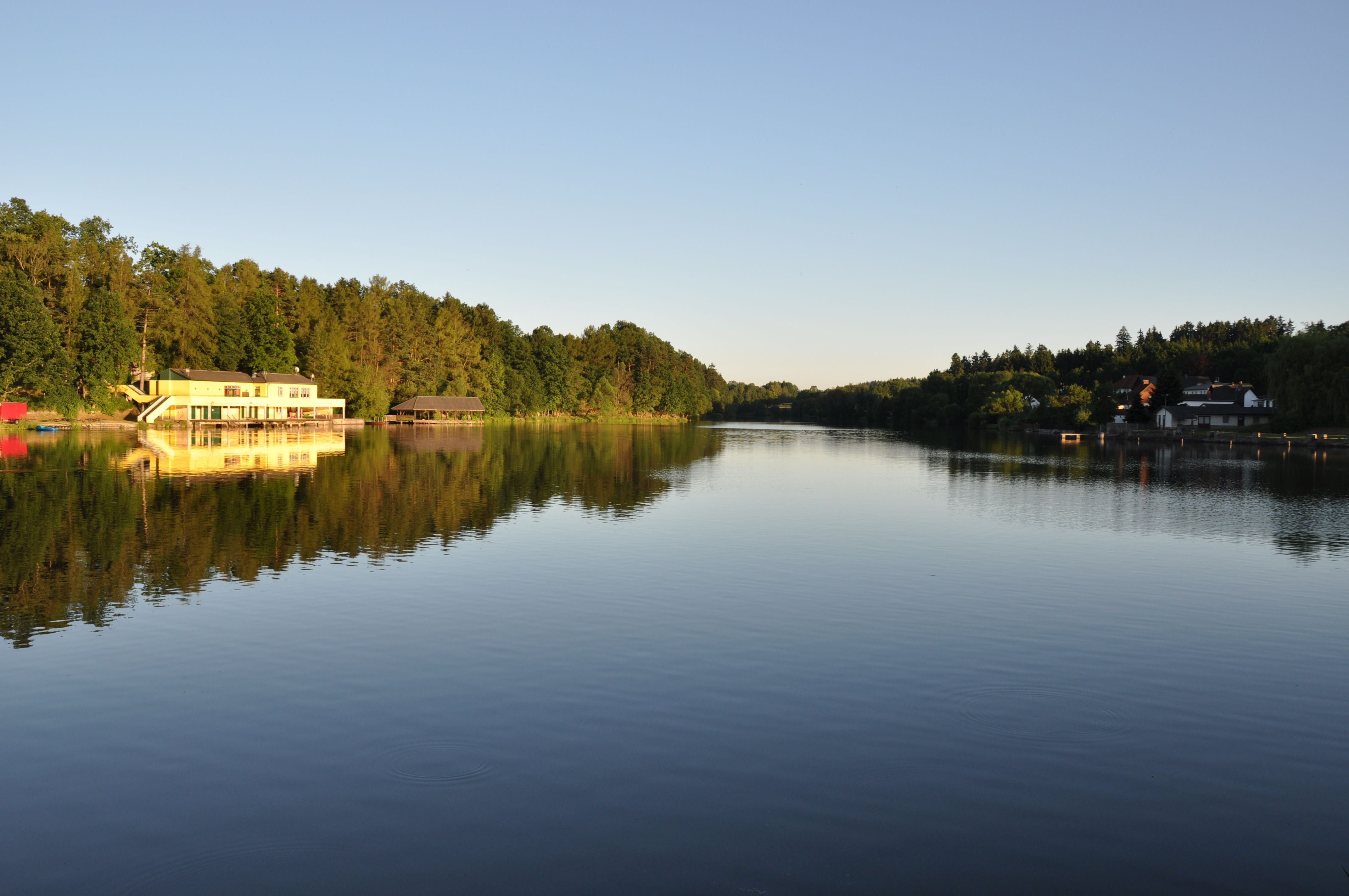 A quiet lake with a building on the shore, surrounded by trees, at sunset.