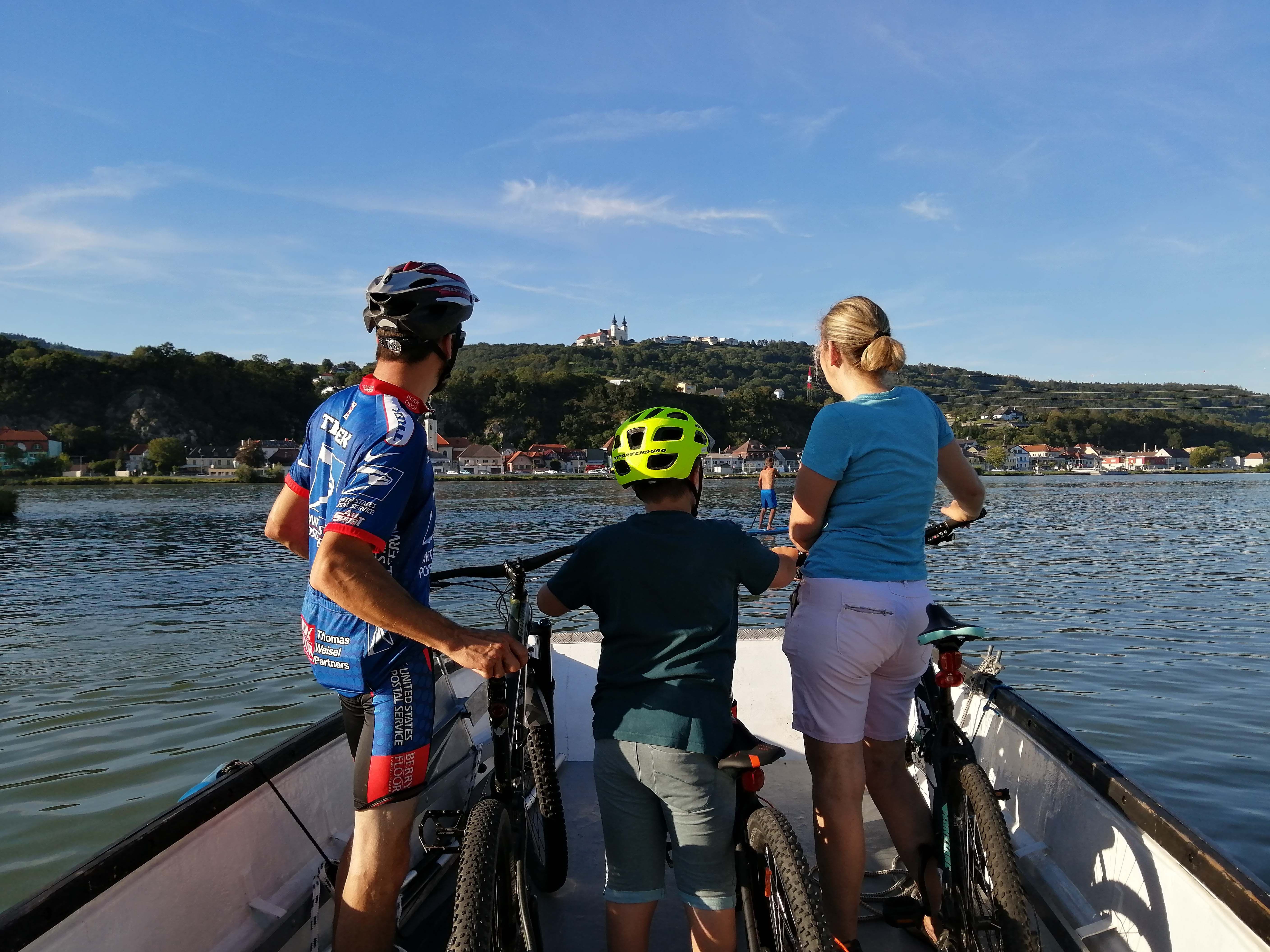 Three people with bicycles on a boat crossing a river. A town and a hill with a building can be seen in the background.