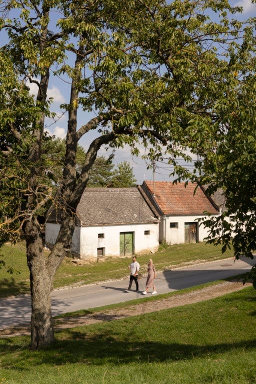 Two people walk past old buildings in a rural setting.