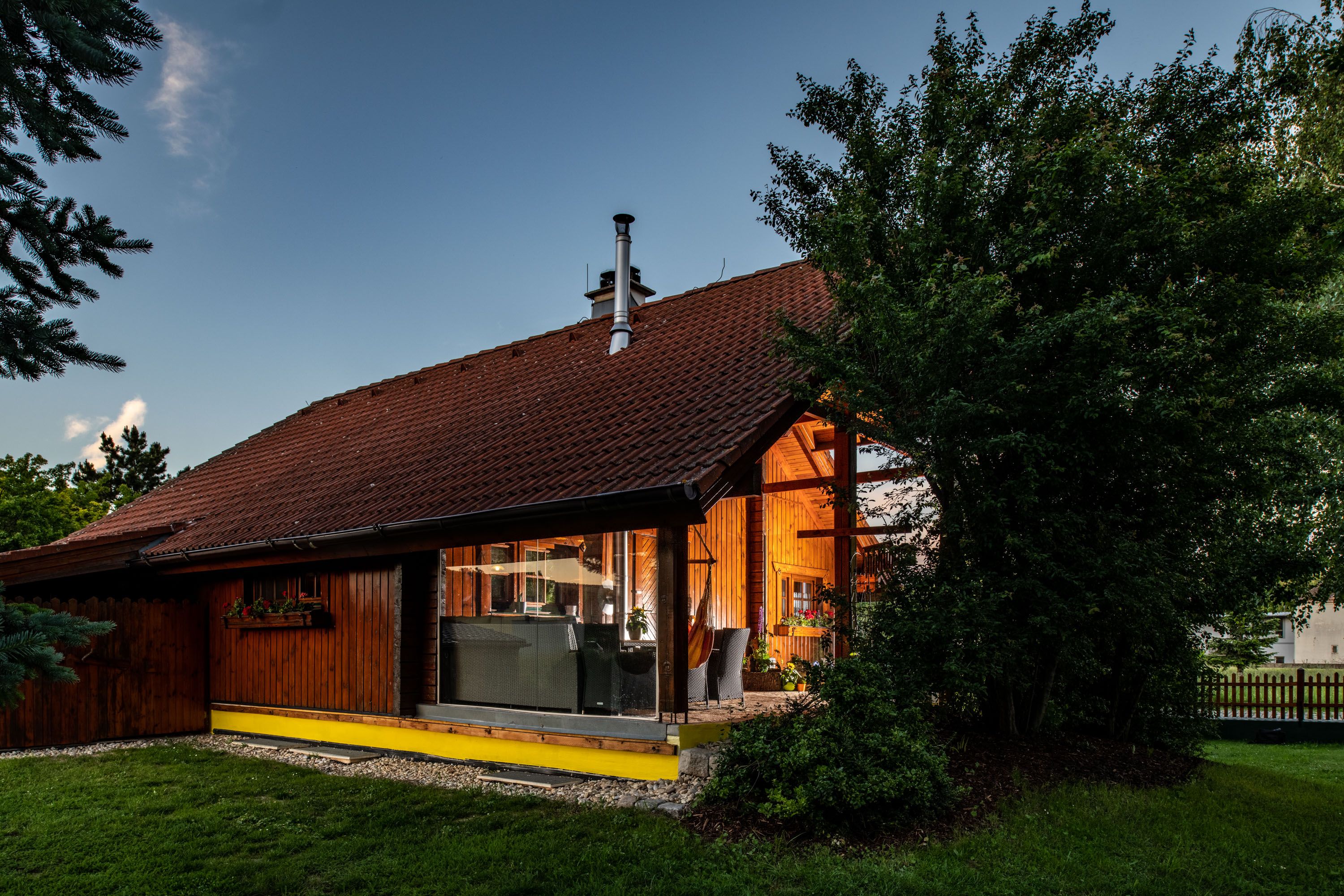 Exterior view of a wooden house with large windows and a garden surrounded by trees.