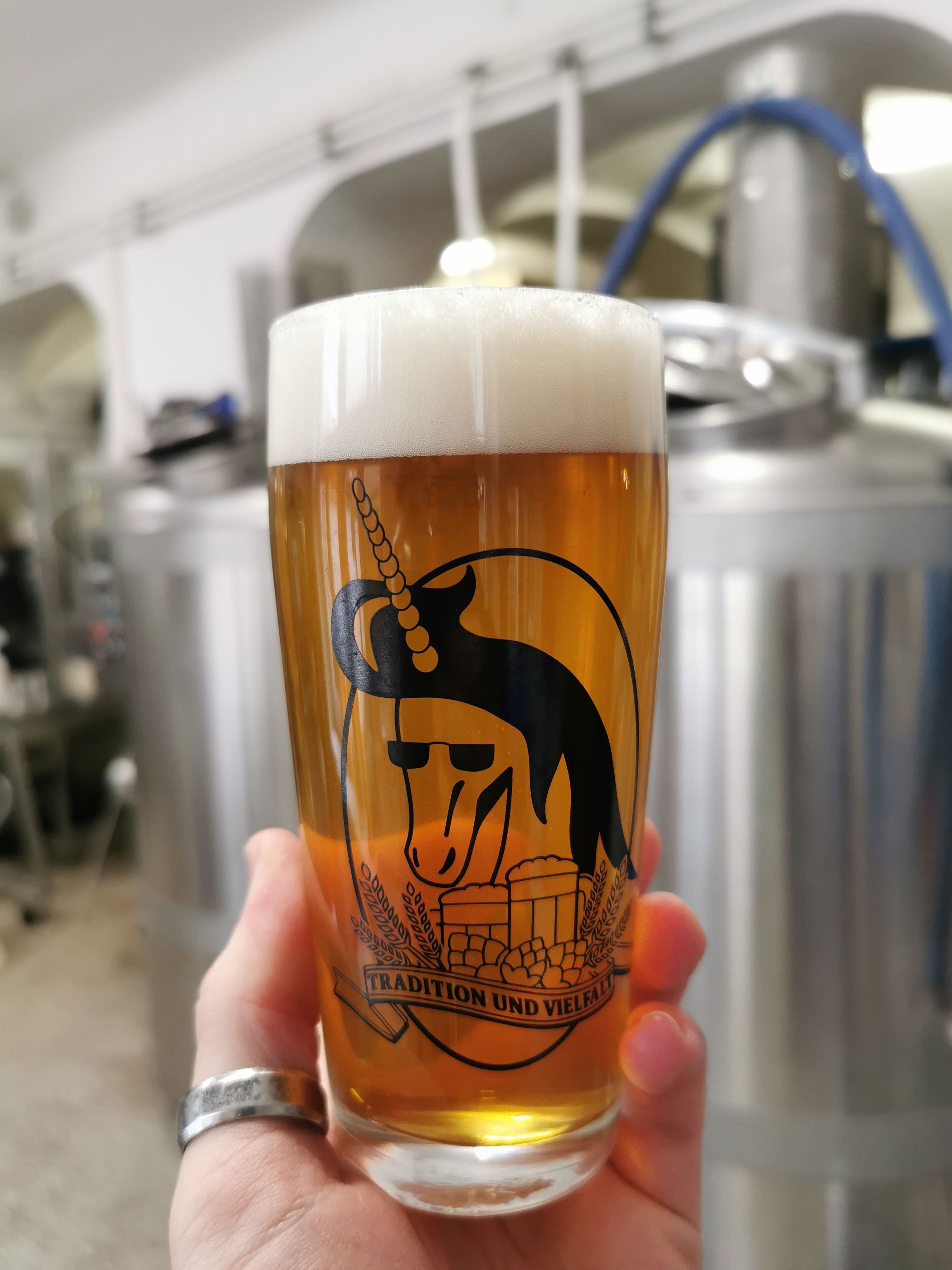 A beer glass with a unicorn logo and the inscription 'Tradition and diversity' in front of brewery equipment.