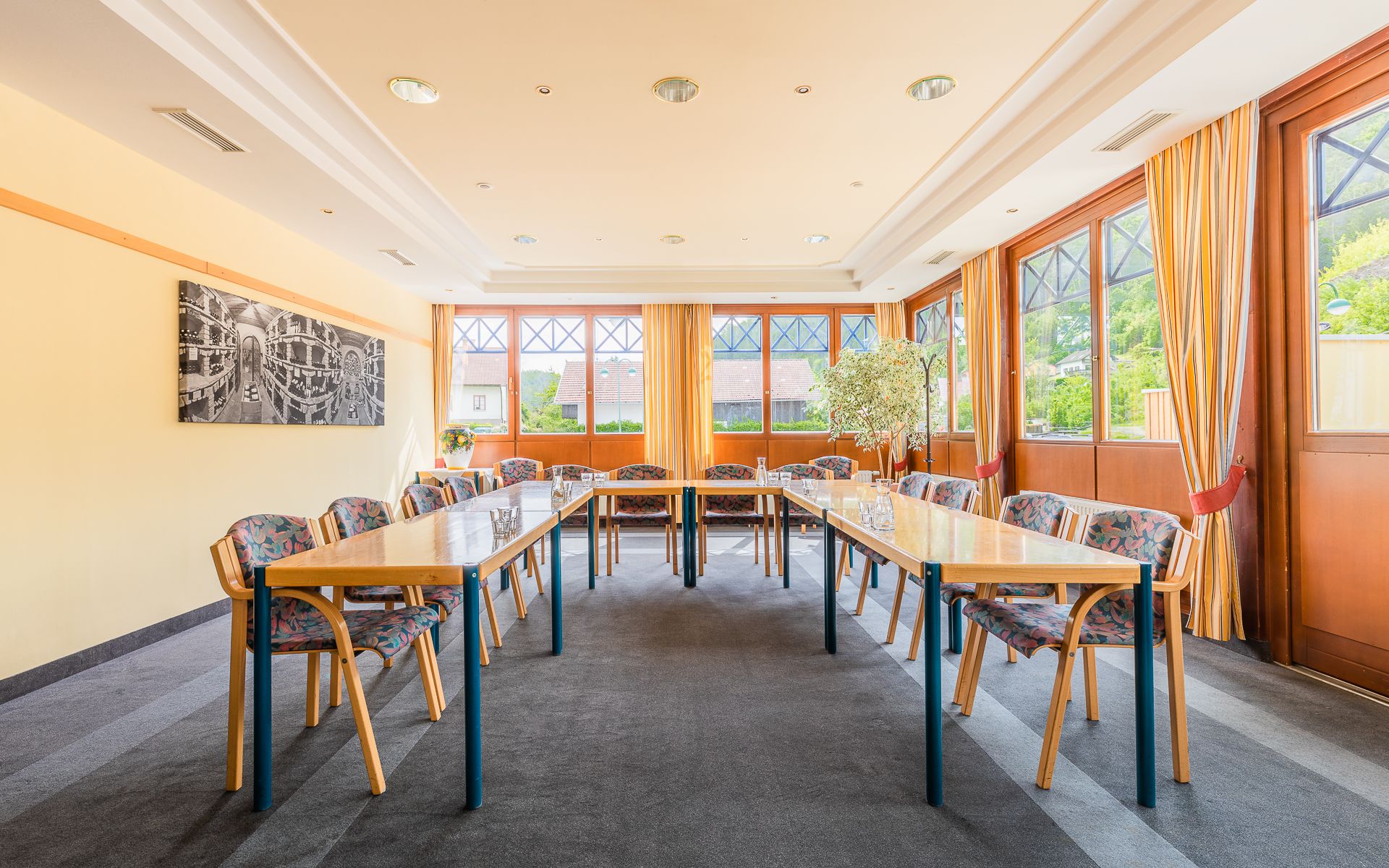 A bright seminar room with U-shaped tables, chairs and large windows with curtains.