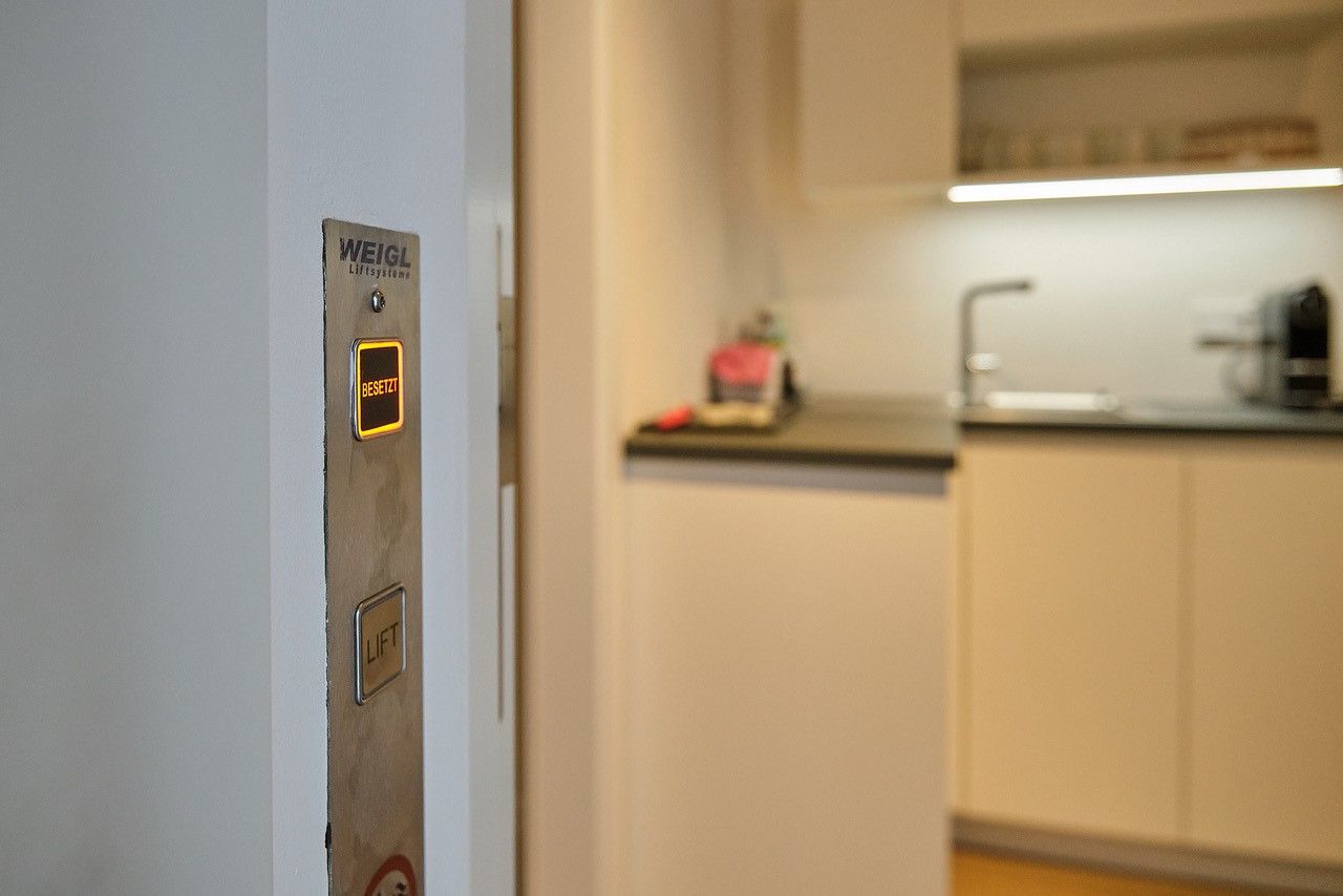 An elevator button with the inscription 'Busy' lights up in a modern living room next to a kitchen.