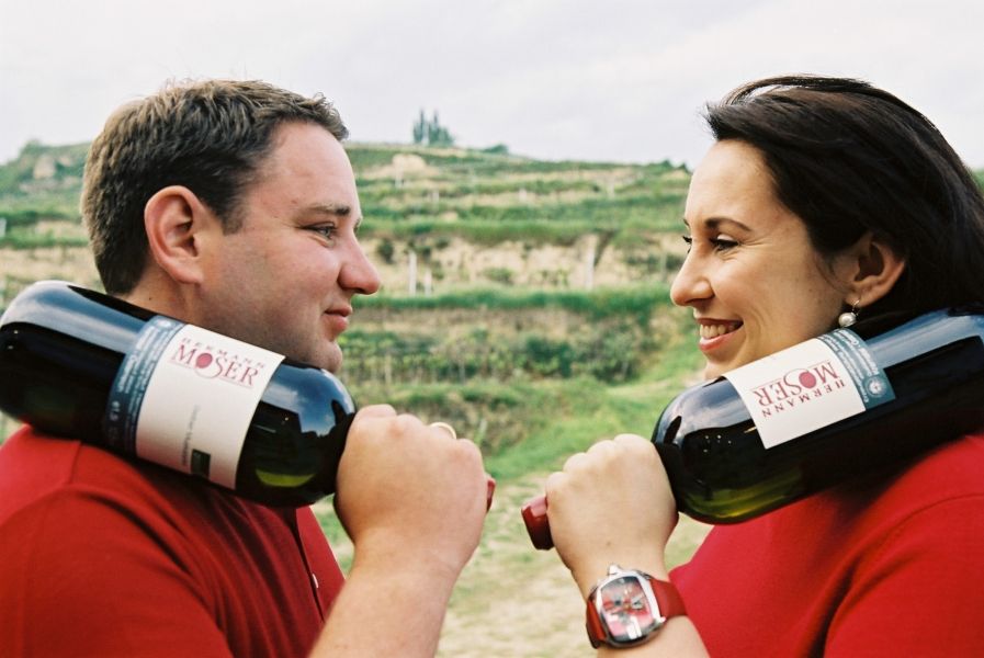 Two people with wine bottles on their shoulders in a vineyard.