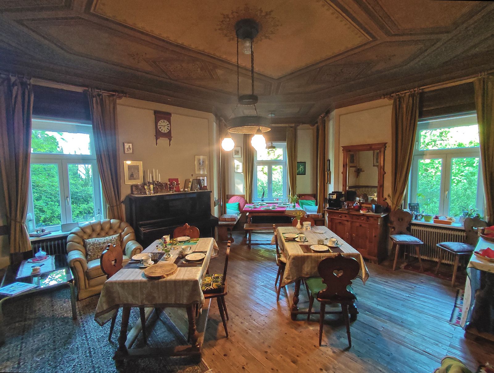 A cozy breakfast room with laid tables, antique furniture and large windows that let in plenty of natural light.