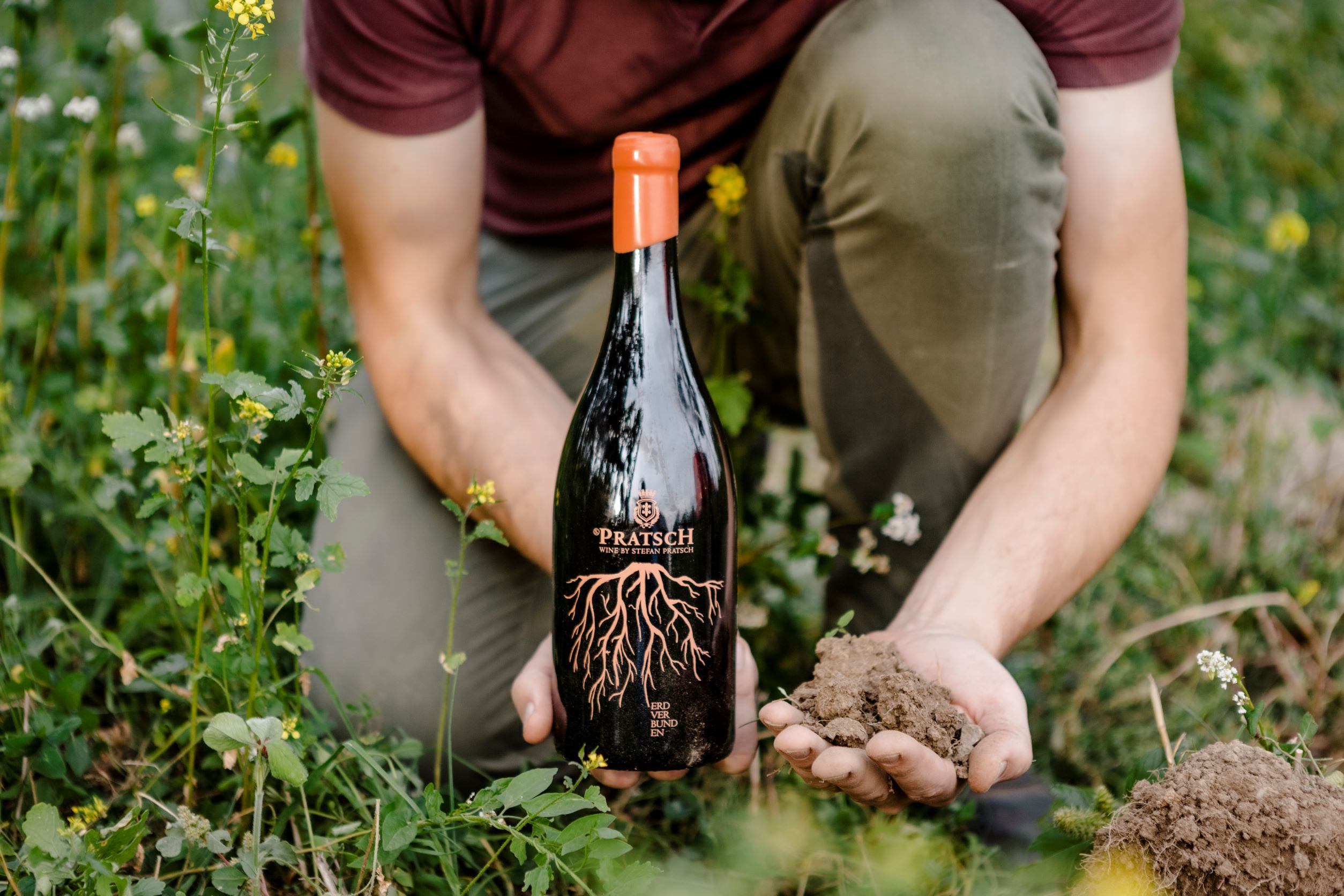 A person holds a bottle of Pratsch wine and a handful of soil in a green field.
