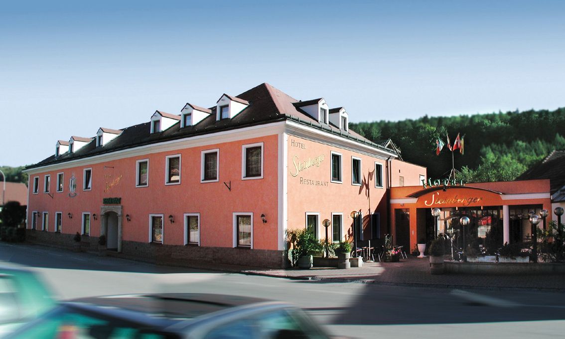 A pink-colored hotel building with several windows and an entrance with a canopy. A car drives past in the foreground.