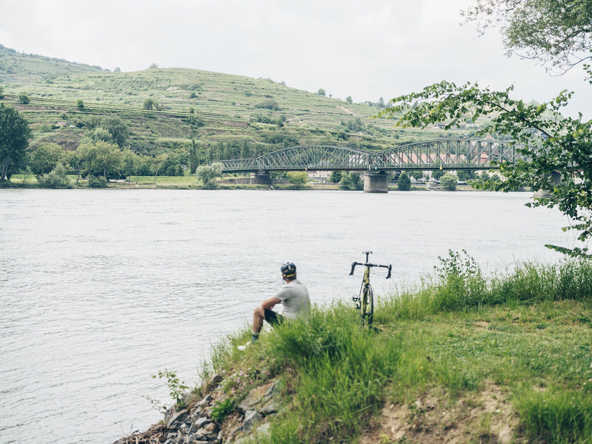 Am Ufer der Donau, umgeben von sanften Hügeln und üppigem Grün, genießt ein Radfahrer die Ruhe der Natur. Die sanften Wellen des Flusses spiegeln die Wolken am Himmel wider und laden zu einer entspannenden Pause ein. Hier, wo die Landschaft atemberaubende Ausblicke bietet, wird jeder Moment zum Erlebnis.