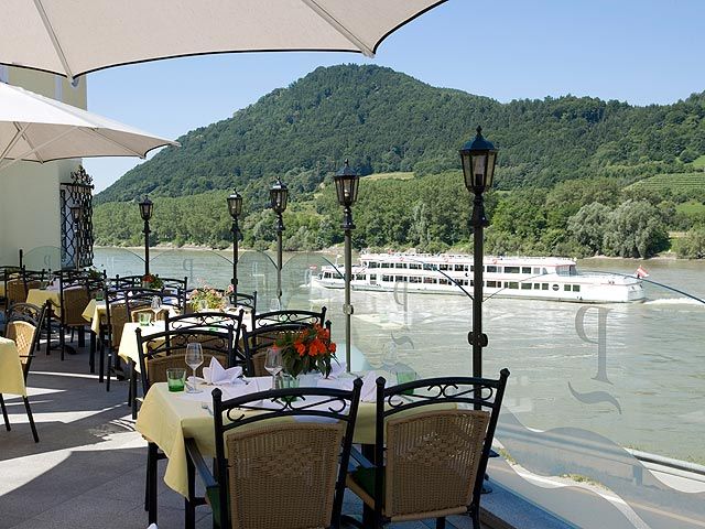 Terrace with covered tables, view of the river and passing ship, surrounded by green hills.