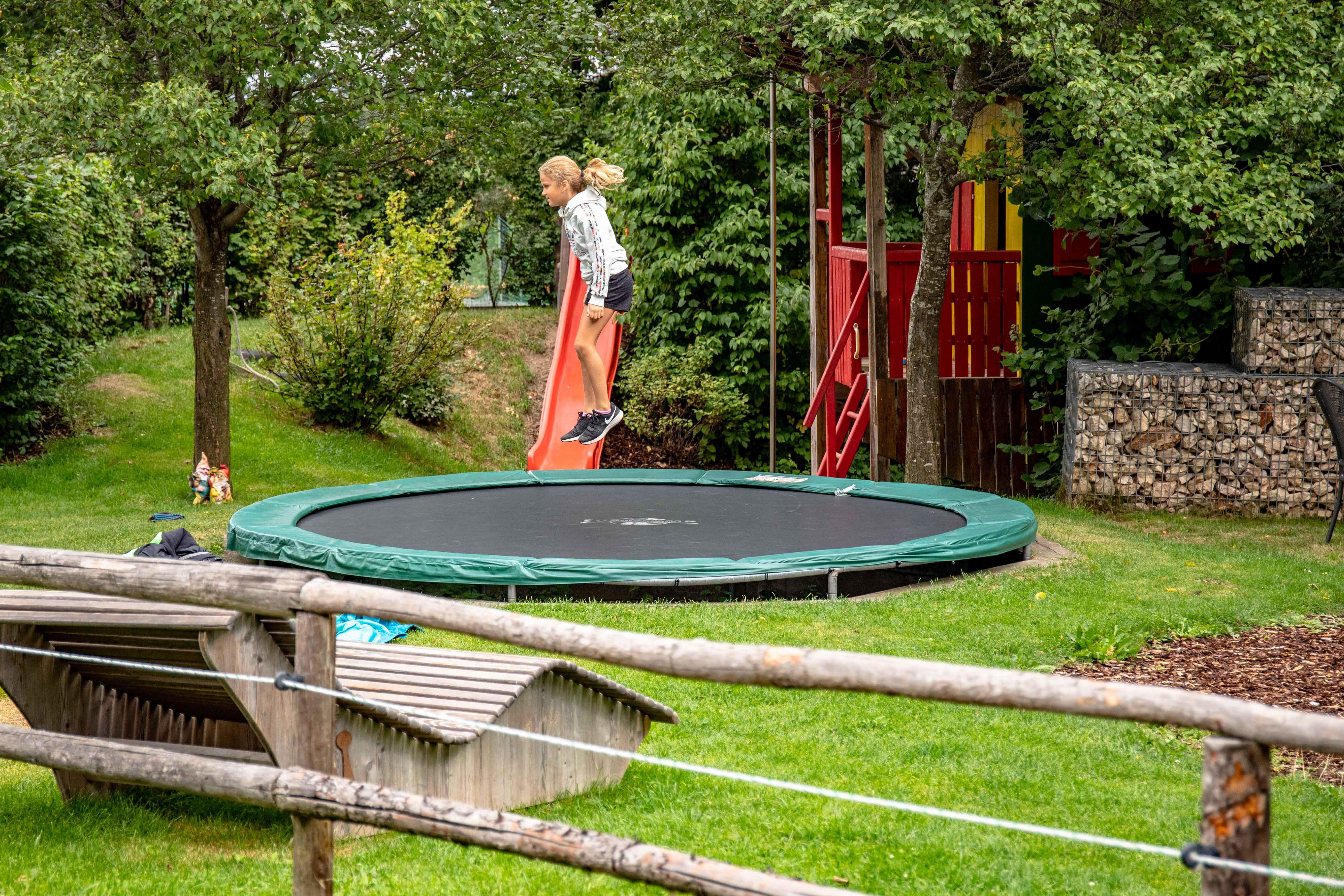 A child jumps on a trampoline in a garden with a slide and playhouse.
