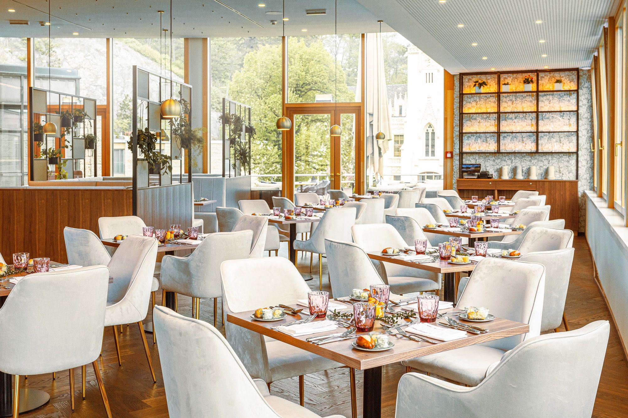 Elegant restaurant with laid tables and white chairs, large windows with a view of the greenery.