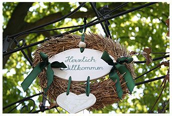 A welcome sign in a wreath with green ribbons, hung on a metal frame, against a background of green leaves.
