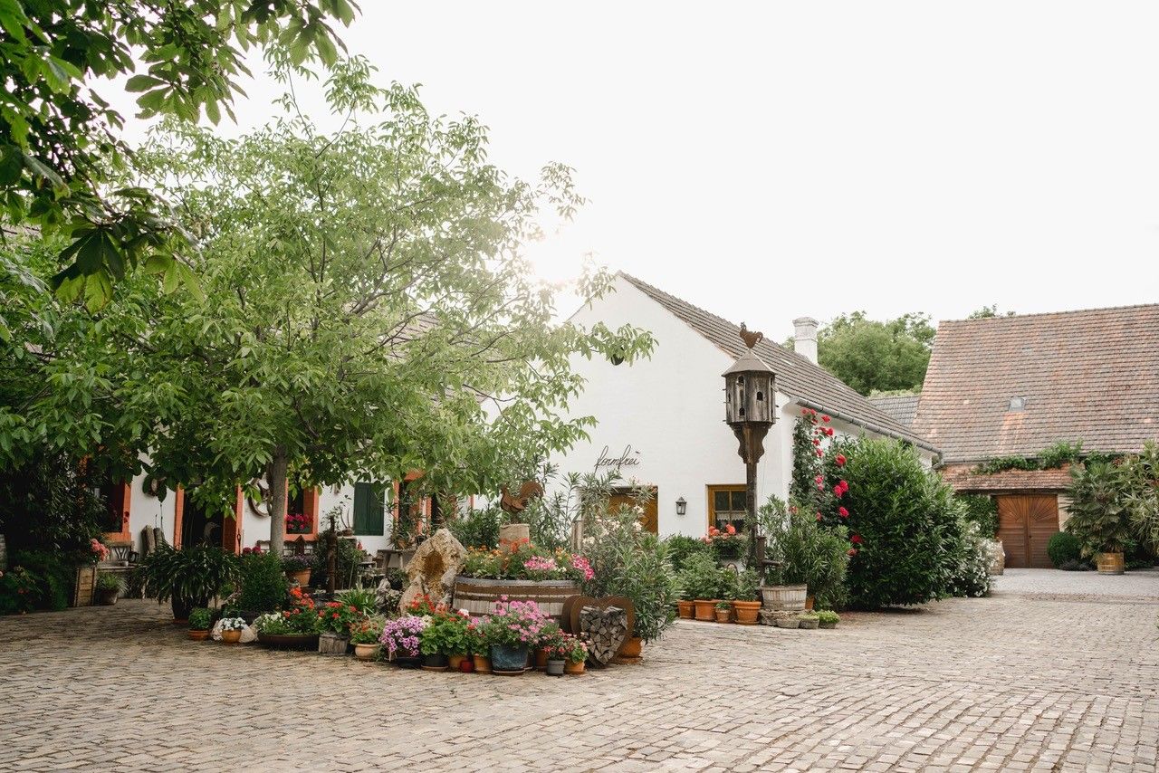 An idyllic courtyard with cobblestones, surrounded by green trees and colorful flowers, in front of a white building with a tiled roof.