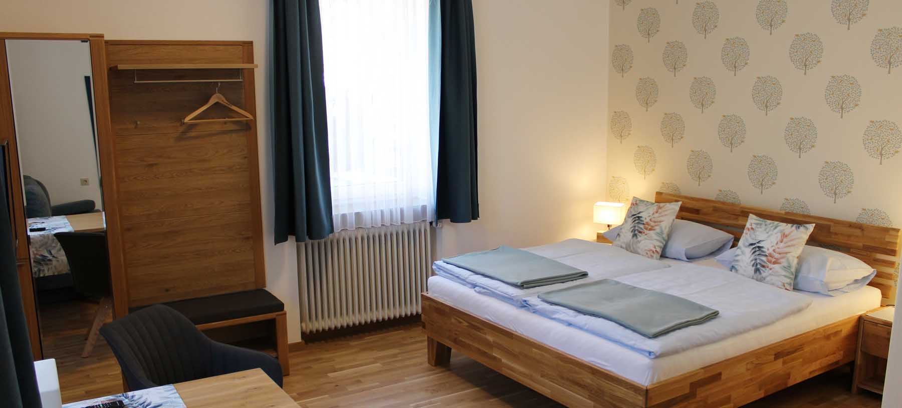 Comfortable double room with bed, closet, table, chair and window with blue curtains.