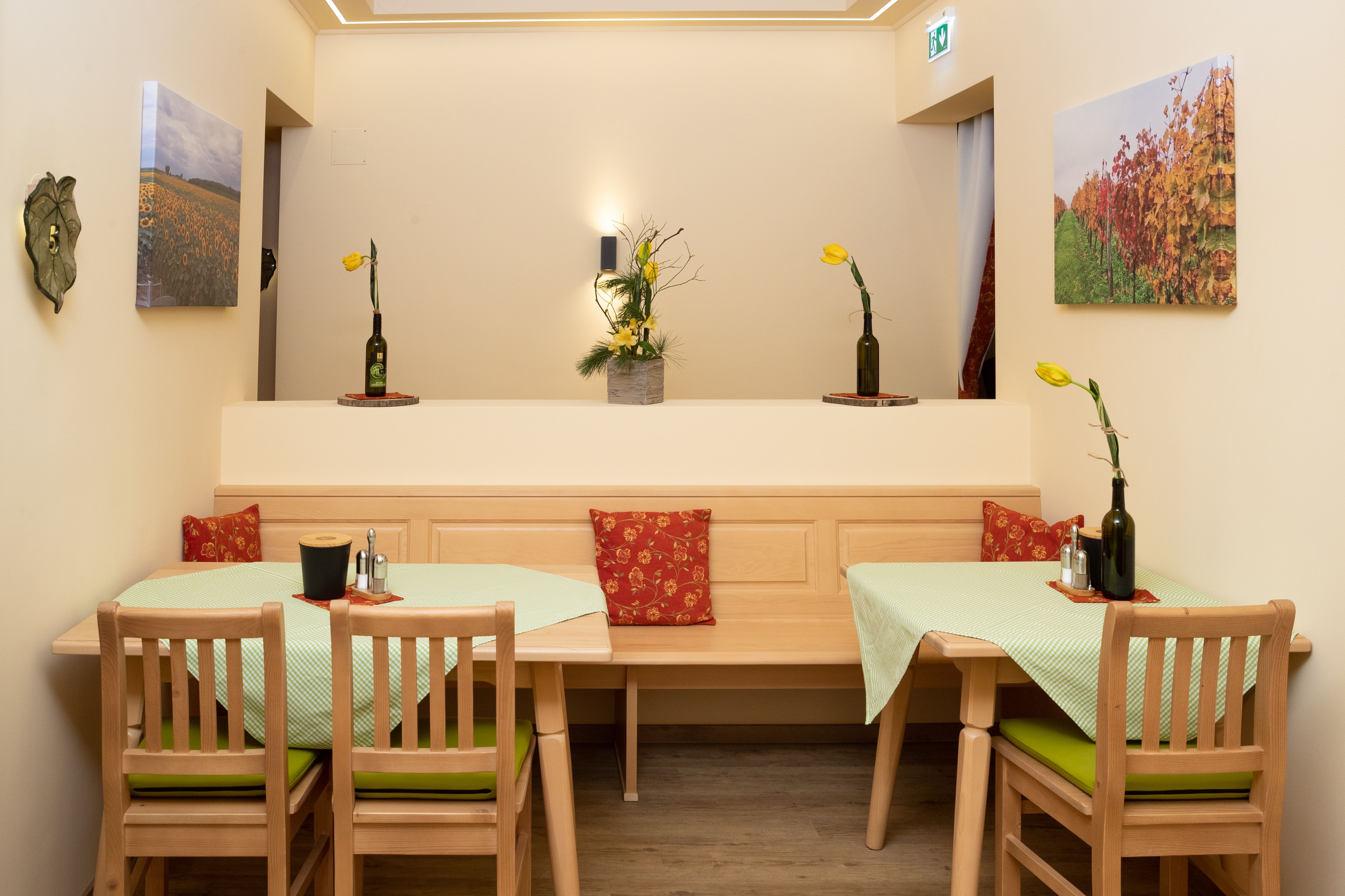 A cozy breakfast room with wooden furniture, green tablecloths and yellow flowers in bottle vases. Landscape paintings hang on the walls.