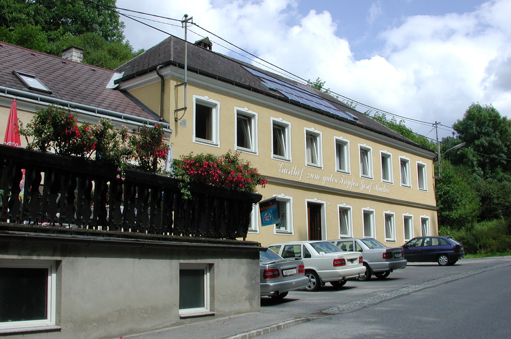 Yellow building with the inscription 'Gasthof zum guten Tropfen Josef Rucker', cars parked in front of it, blue sky in the background.