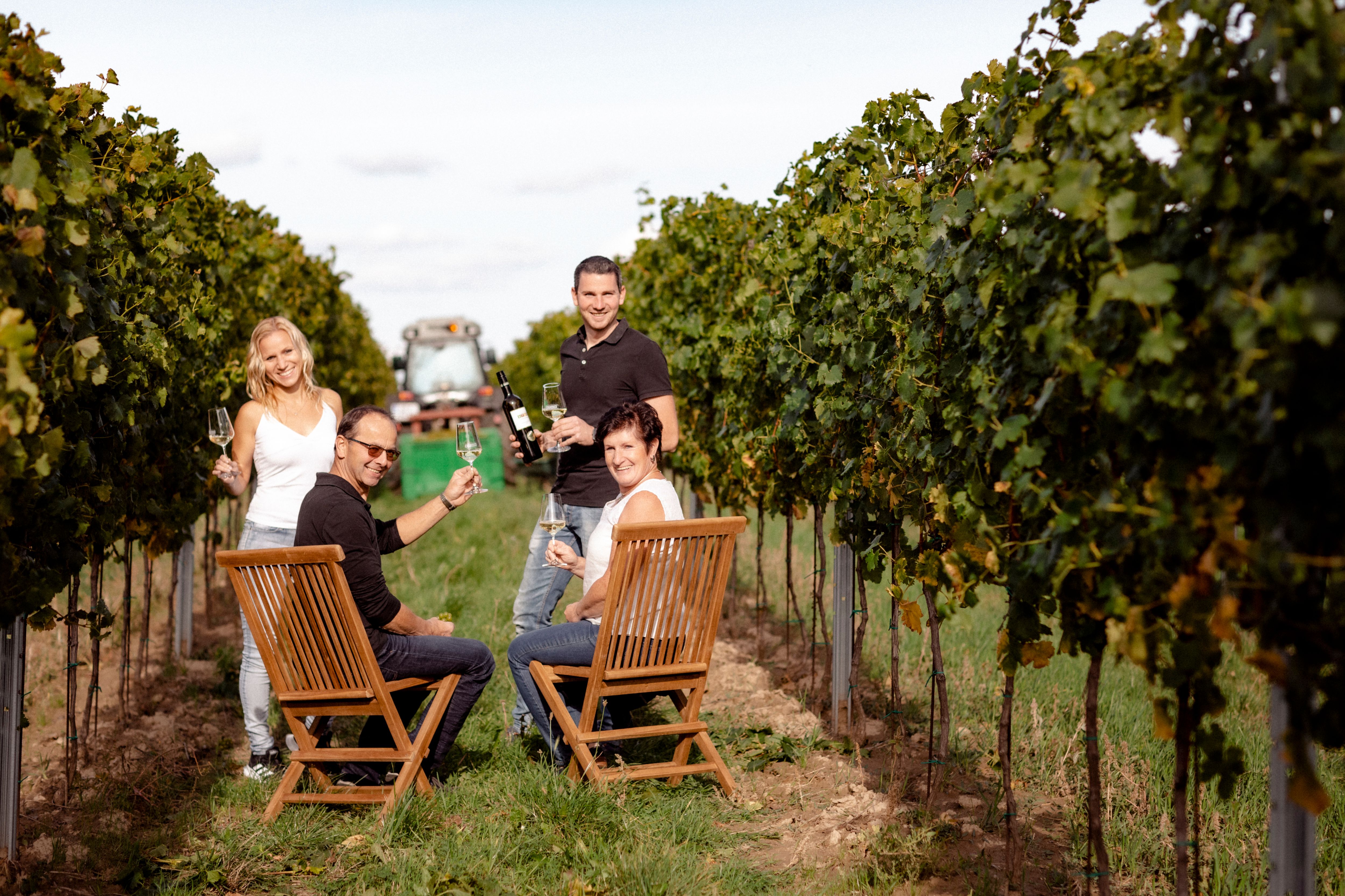 Four people sit and stand in a vineyard with wine glasses in their hands.