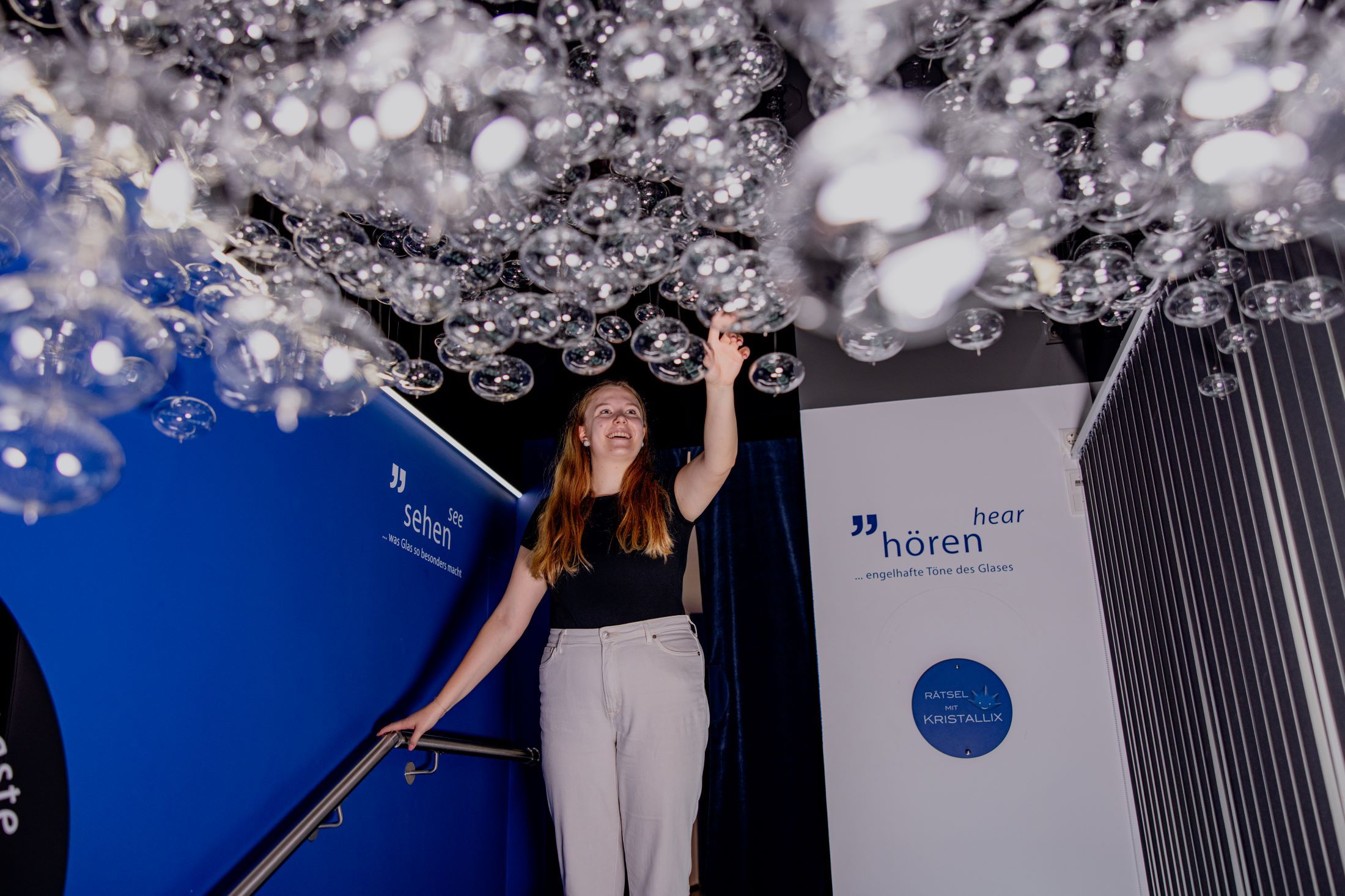 A woman touches hanging glass bubbles in an exhibition with blue walls and inscriptions.