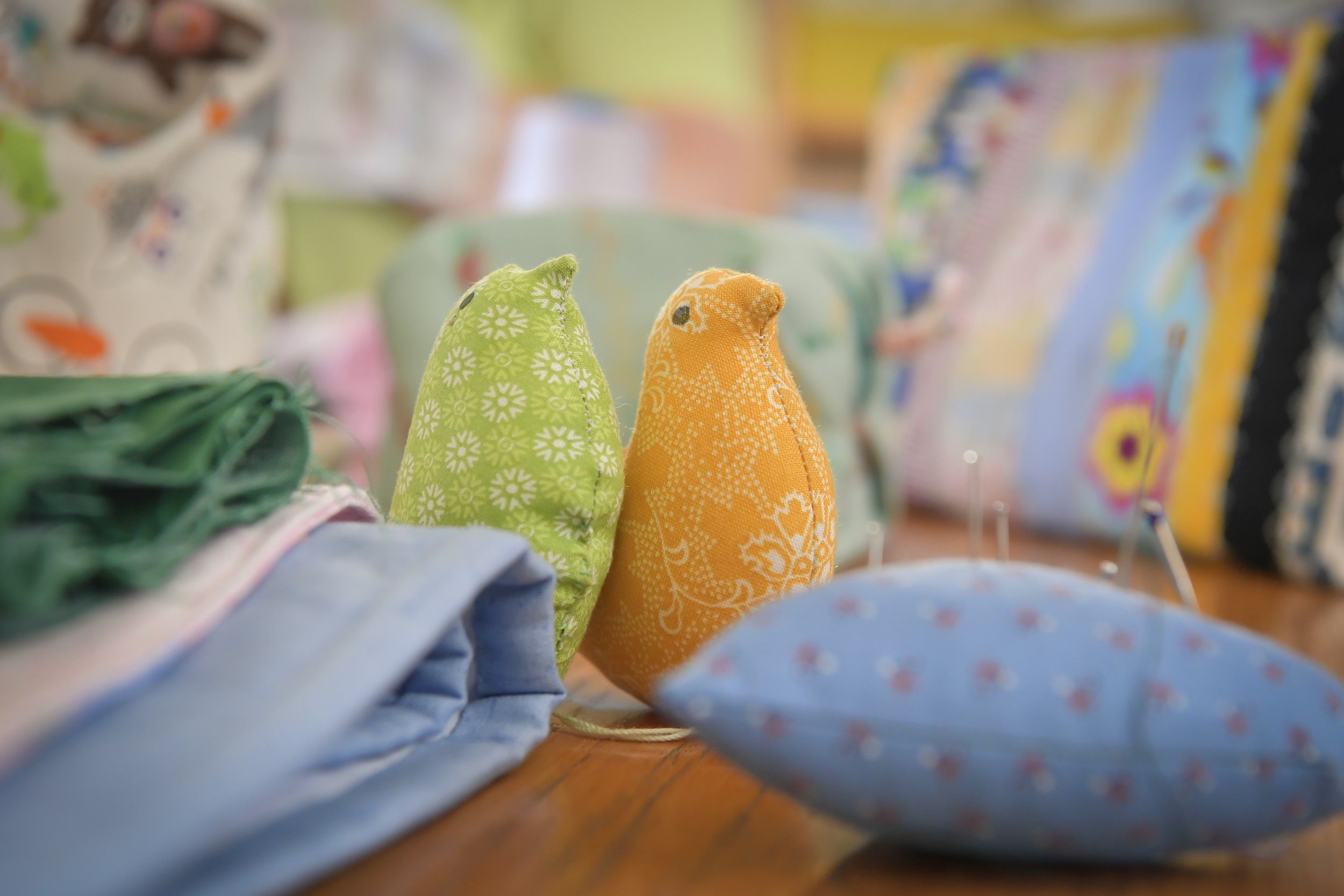 Two fabric birds, one green and one orange, on a table with fabrics and sewing utensils.