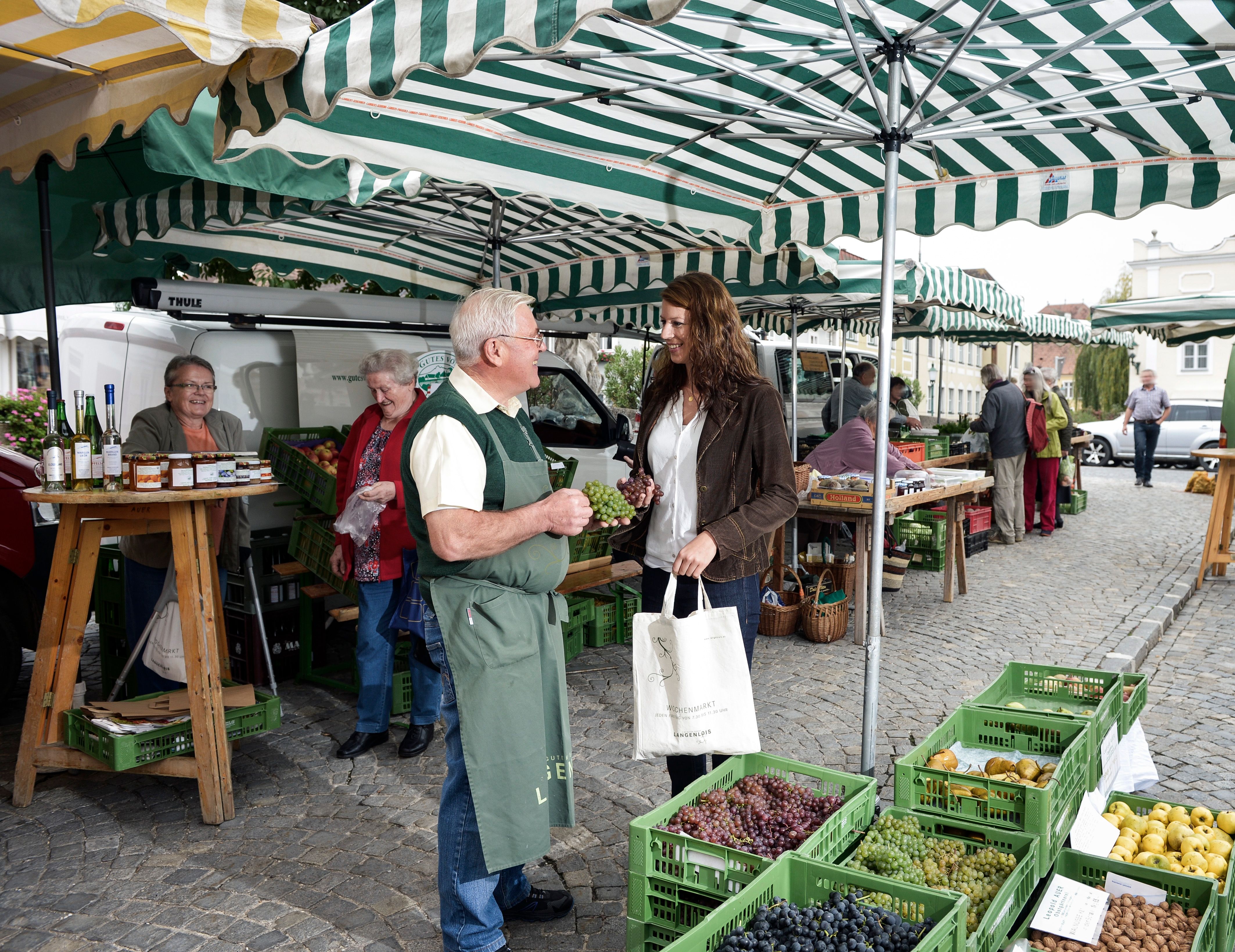 People at a weekly market with fruit and vegetable stalls under striped awnings.