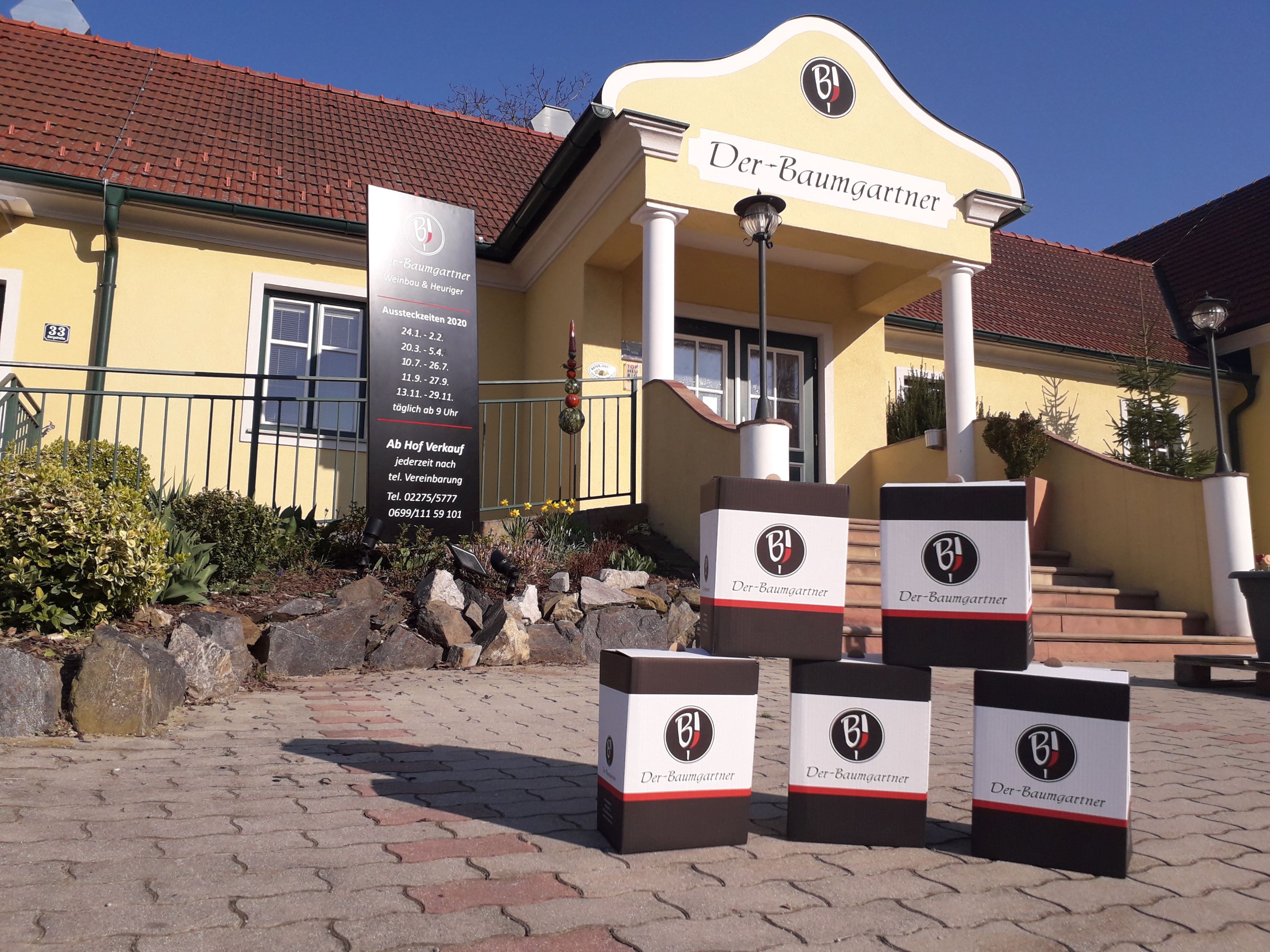 Exterior view of a yellow building with the inscription 'Der-Baumgartner'. In front of the entrance are cardboard boxes with the logo 'BJ'.