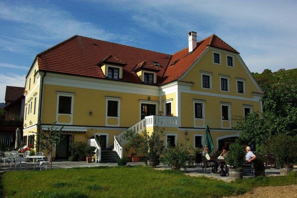 Yellow building with red roof and terrace, surrounded by trees.