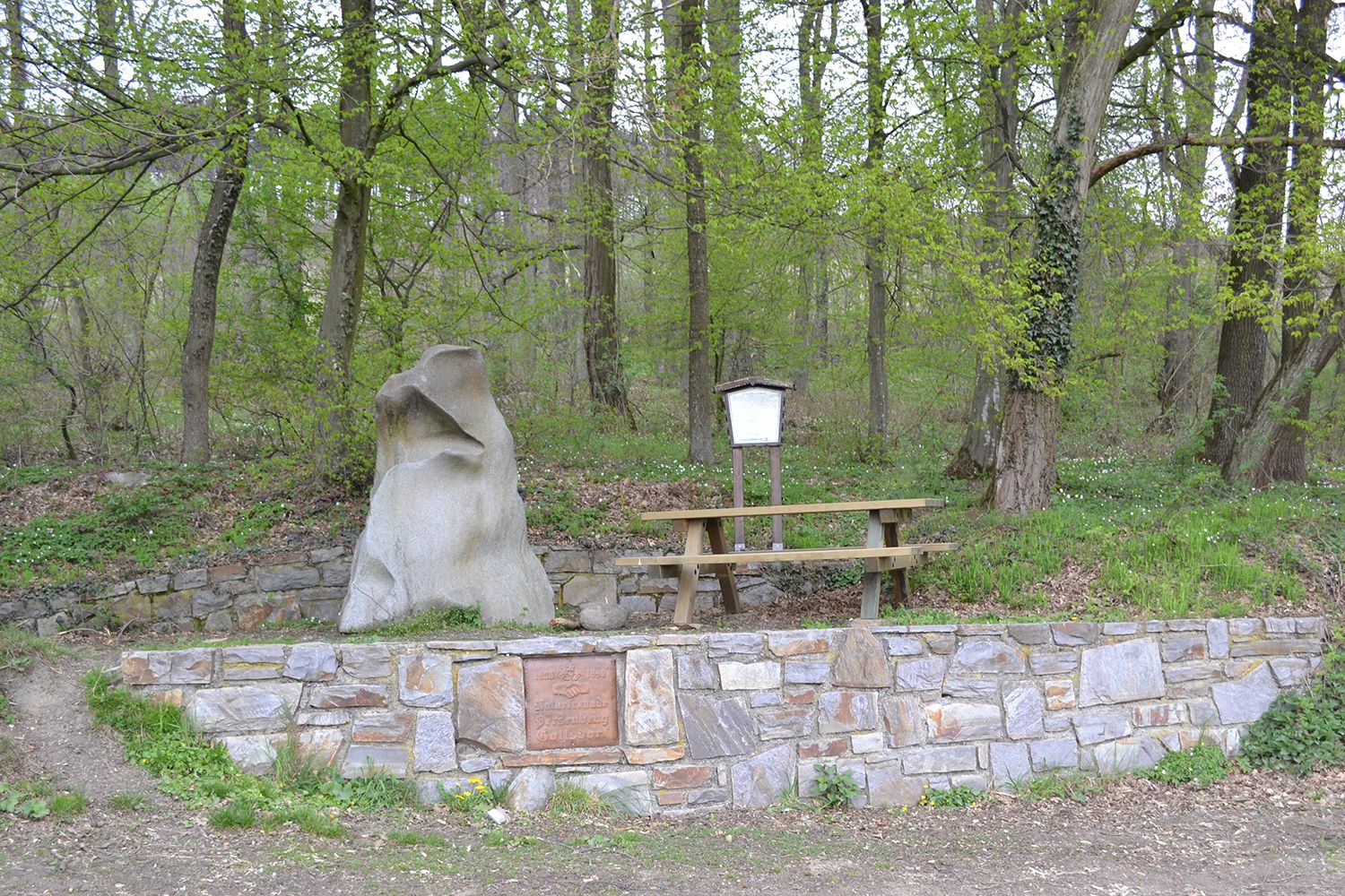 A natural stone and a bench in the forest, surrounded by trees and a stone wall.