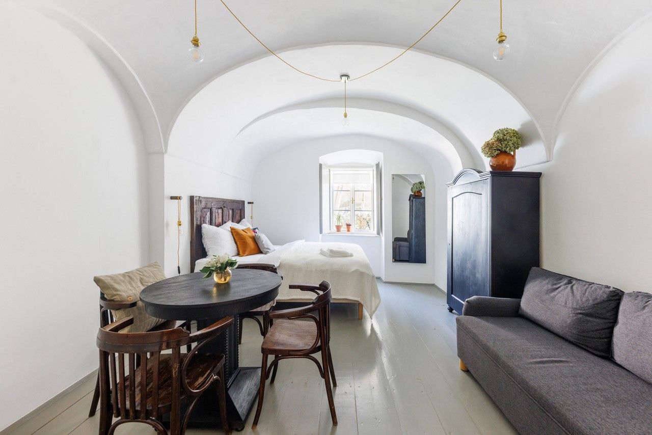 A stylishly furnished guest room with vaulted ceiling, bed, sofa, table and chairs. A window lets in natural light.
