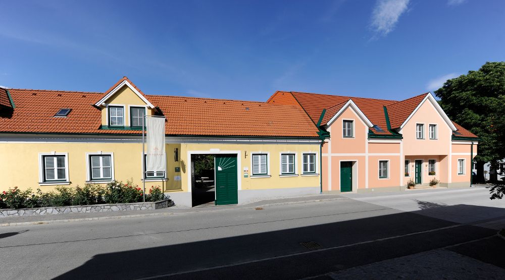 A long, single-storey house with a yellow and pink façade, green shutters and a red tiled roof under a blue sky.
