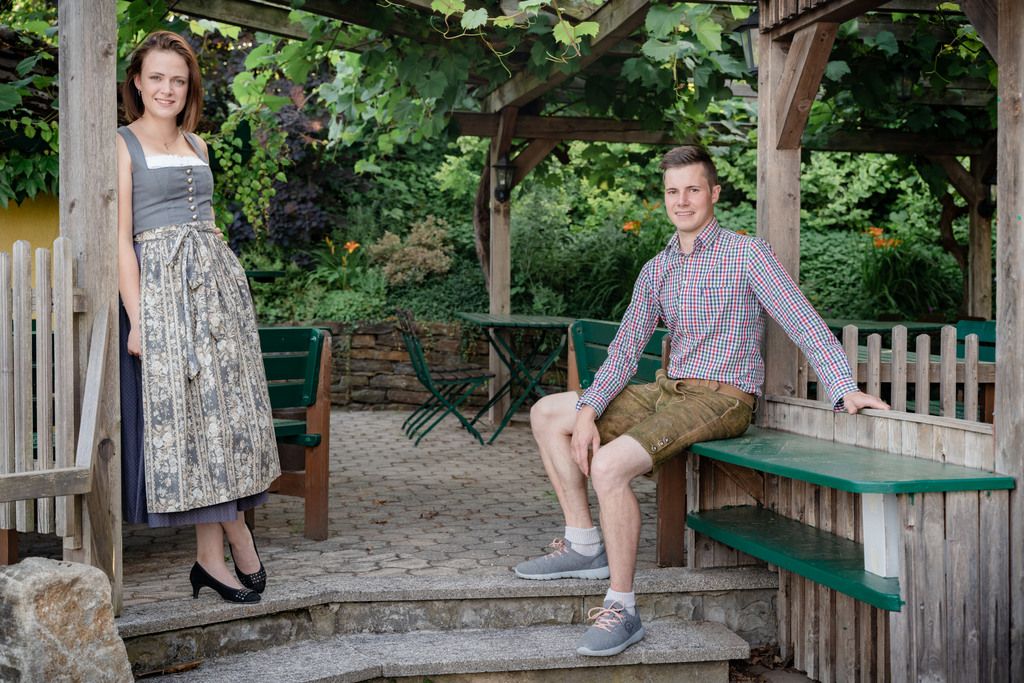 A man in lederhosen and a woman in a dirndl pose in a garden with wooden constructions.