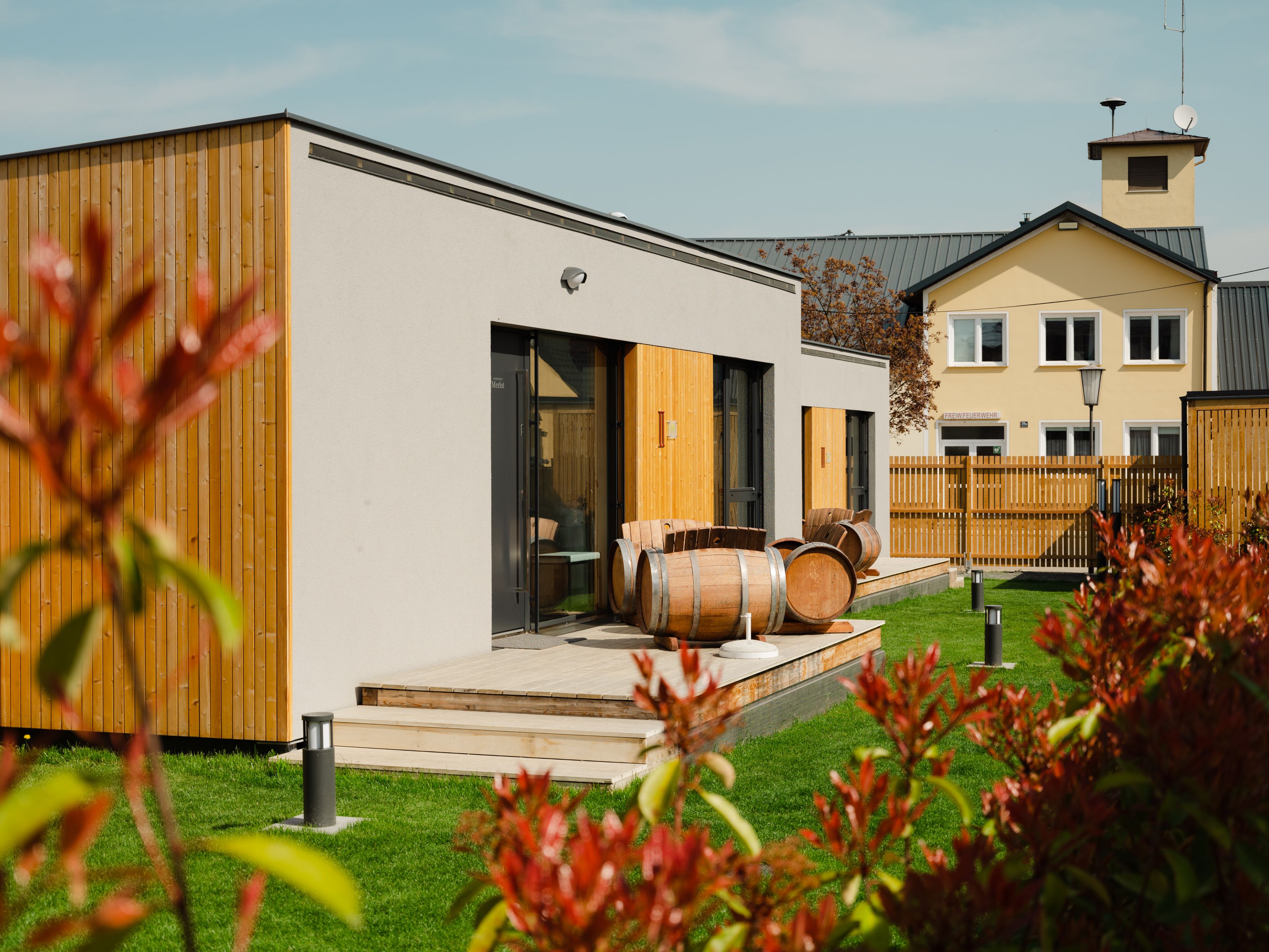 Modern apartments with wood paneling and wine barrels on the terrace, surrounded by green lawns and a fence.