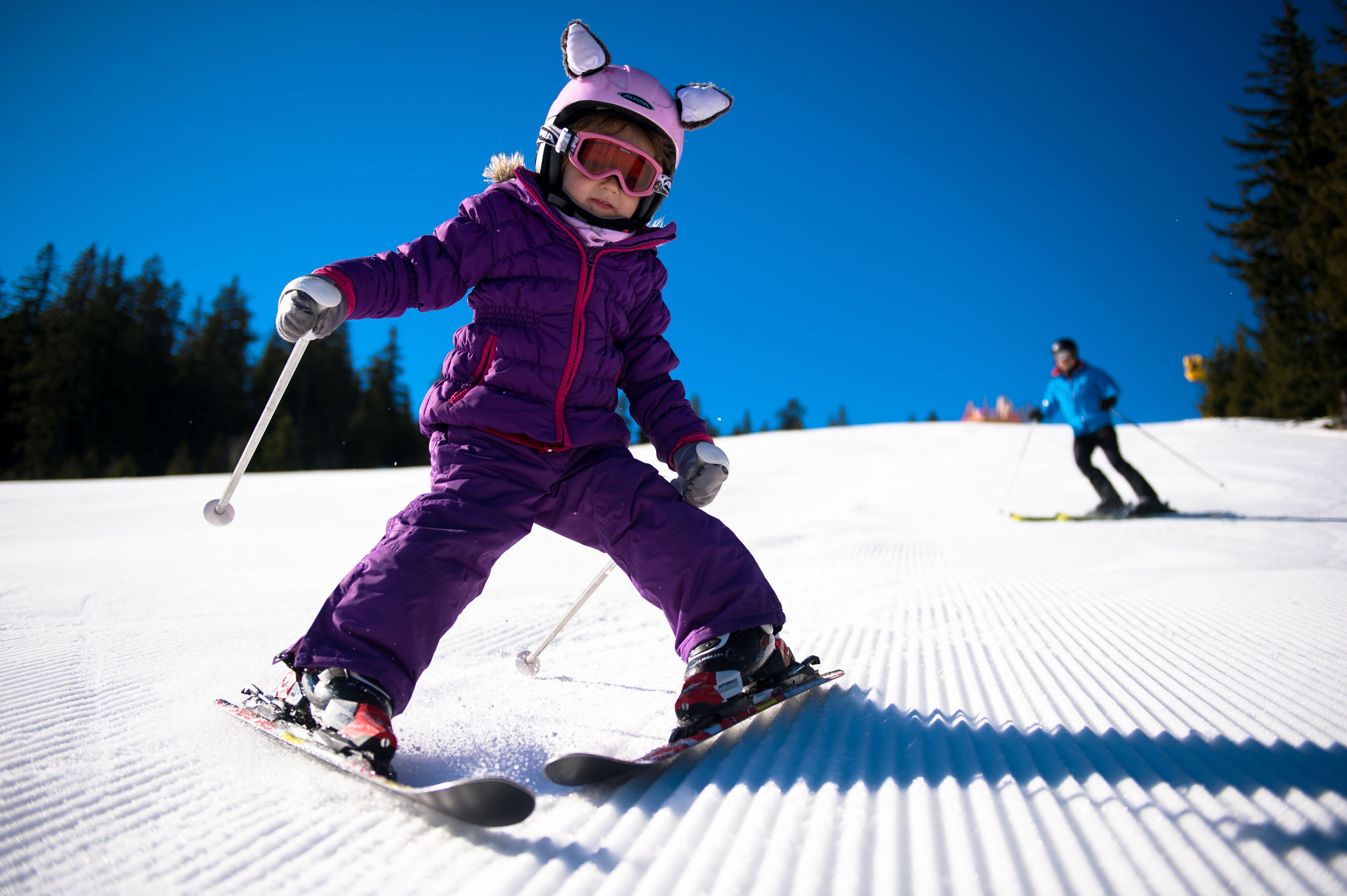 A child in a purple ski suit is skiing on a groomed slope, followed by an adult in a blue suit.