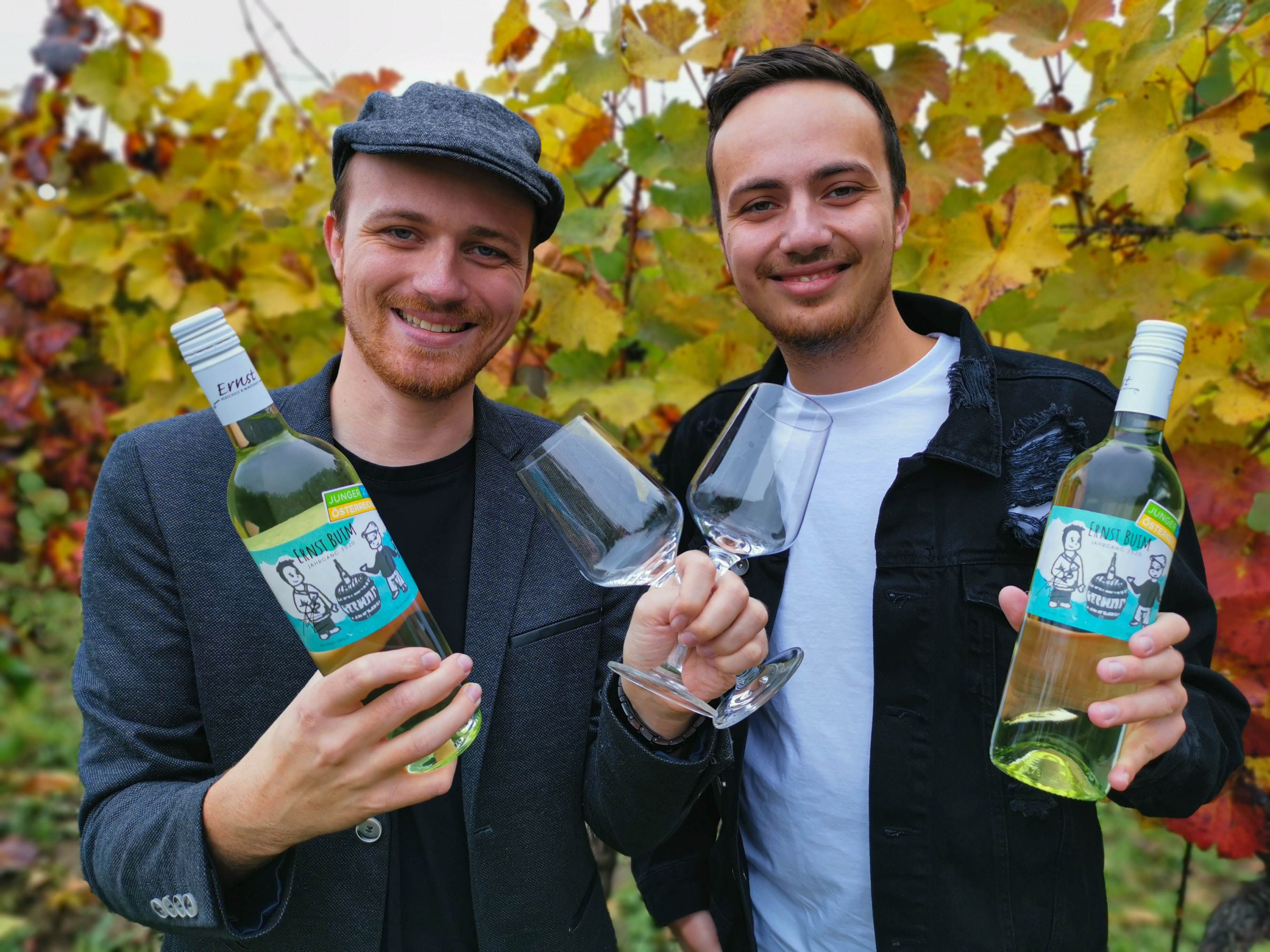 Two men hold wine bottles and glasses in front of autumnal vines.