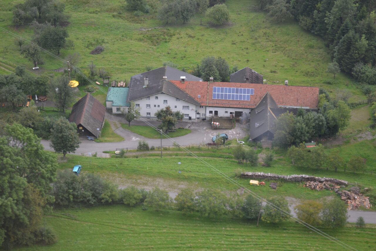 Aerial view of a farm with solar panels on the roof, surrounded by green fields and trees.