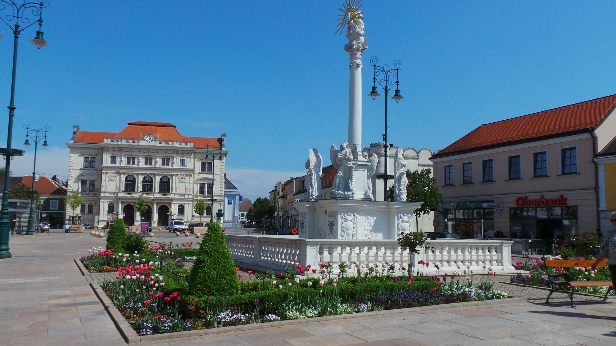 Main square with baroque column and flowerbeds, in the background a historic building.