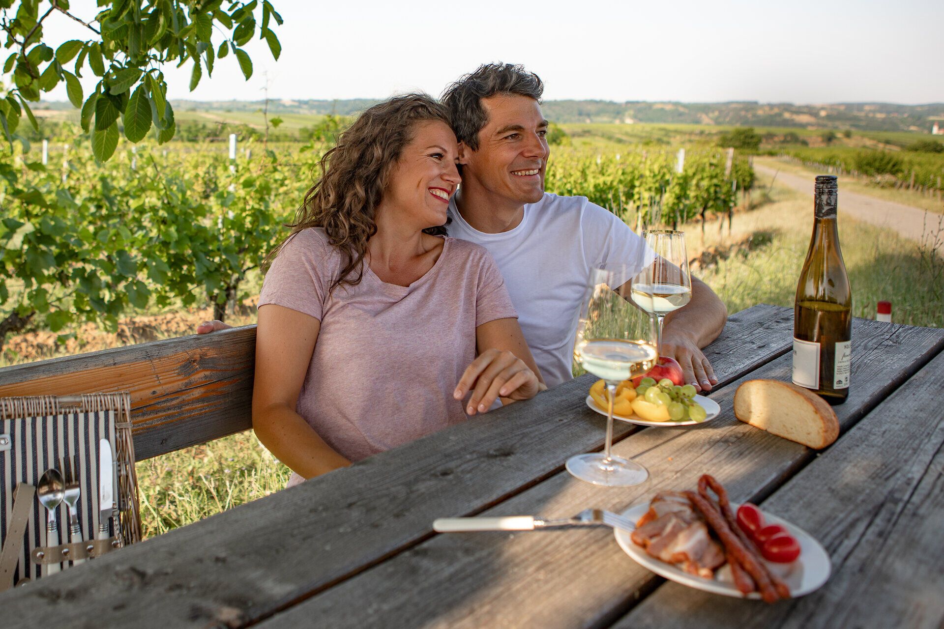 Couple sitting relaxed at a wooden table in the vineyard with bottles of wine.