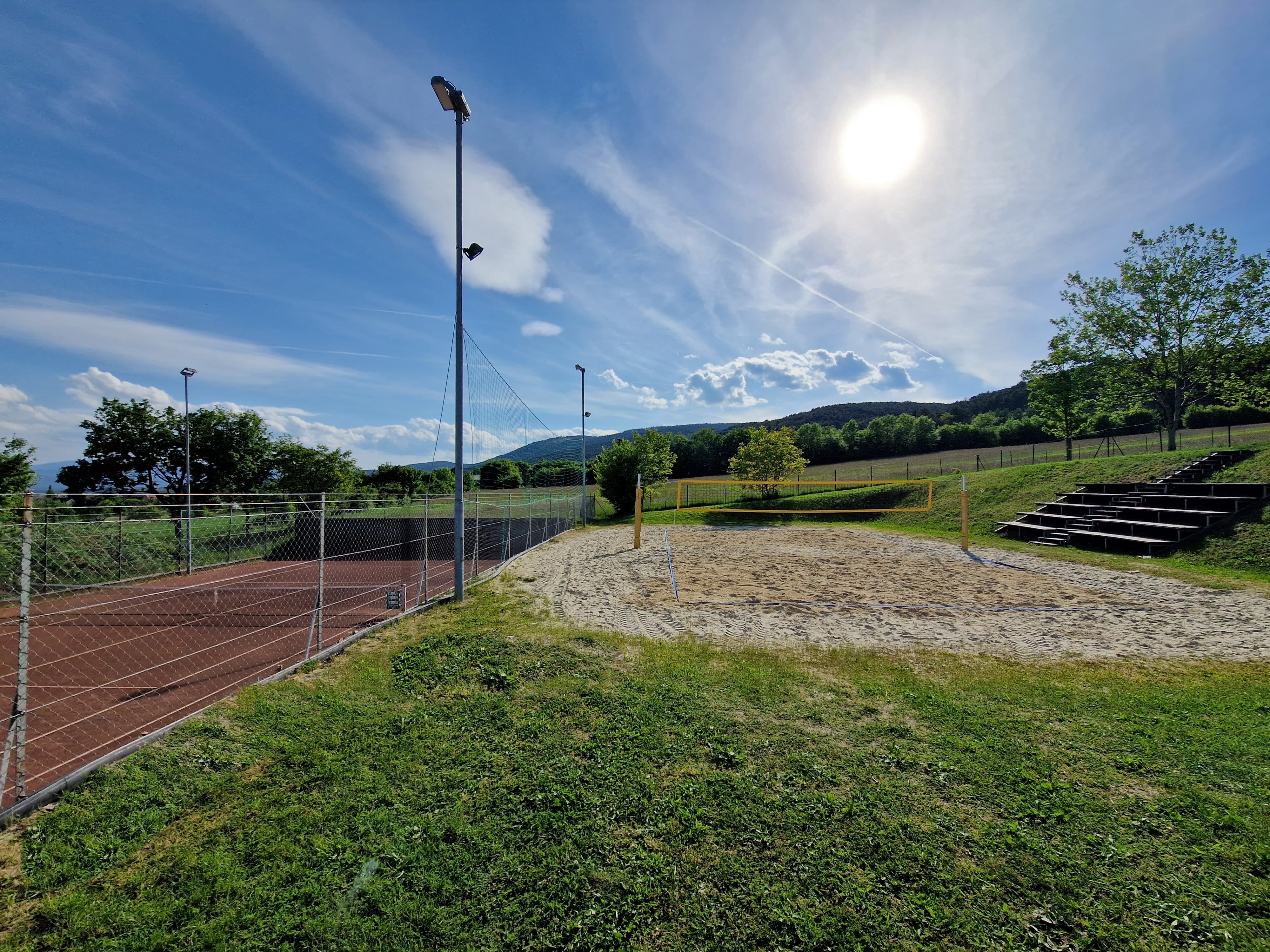 A sunny volleyball court with sand and a net, surrounded by green landscape and trees.