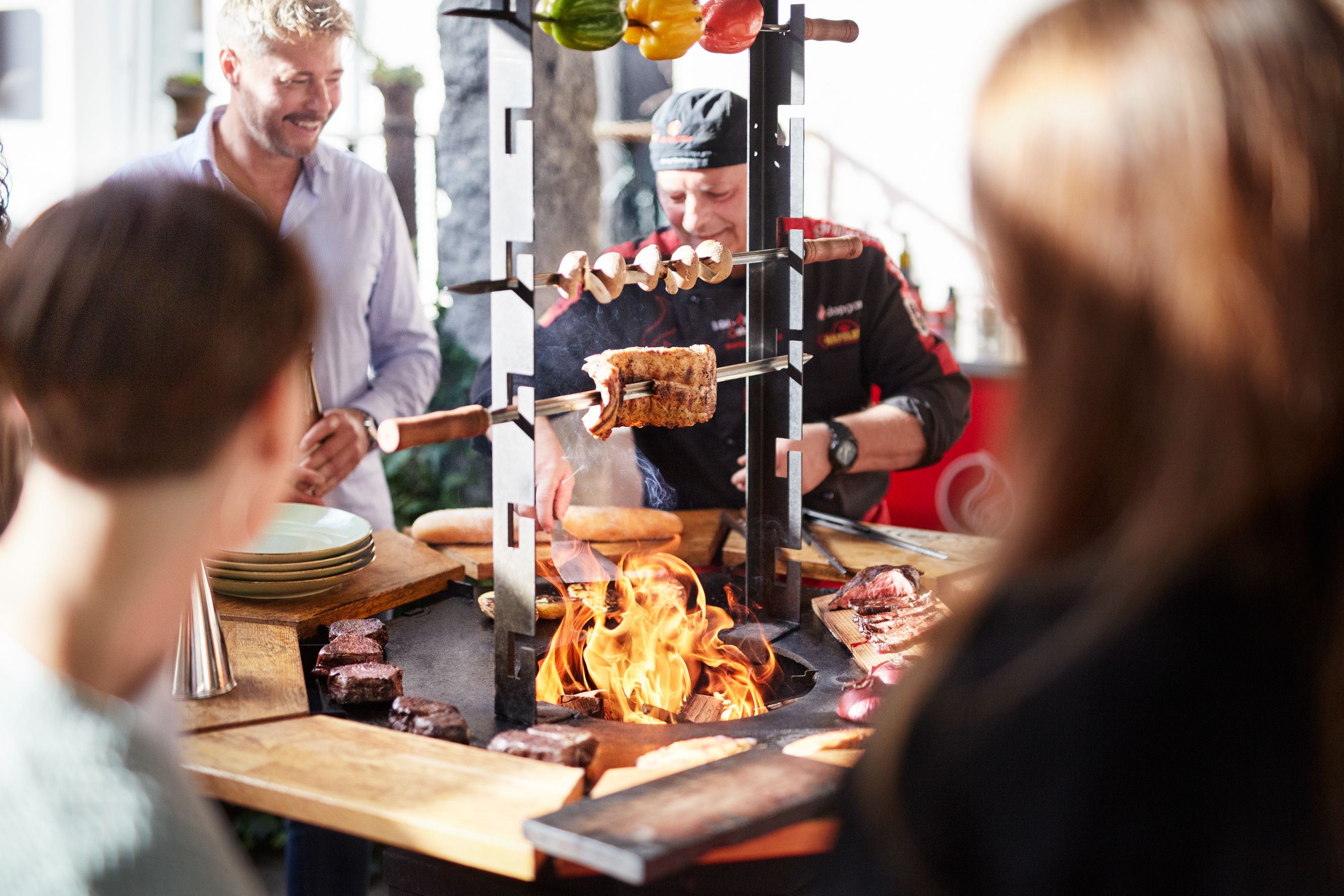 A grill master prepares meat and vegetables on an open grill, surrounded by interested spectators.