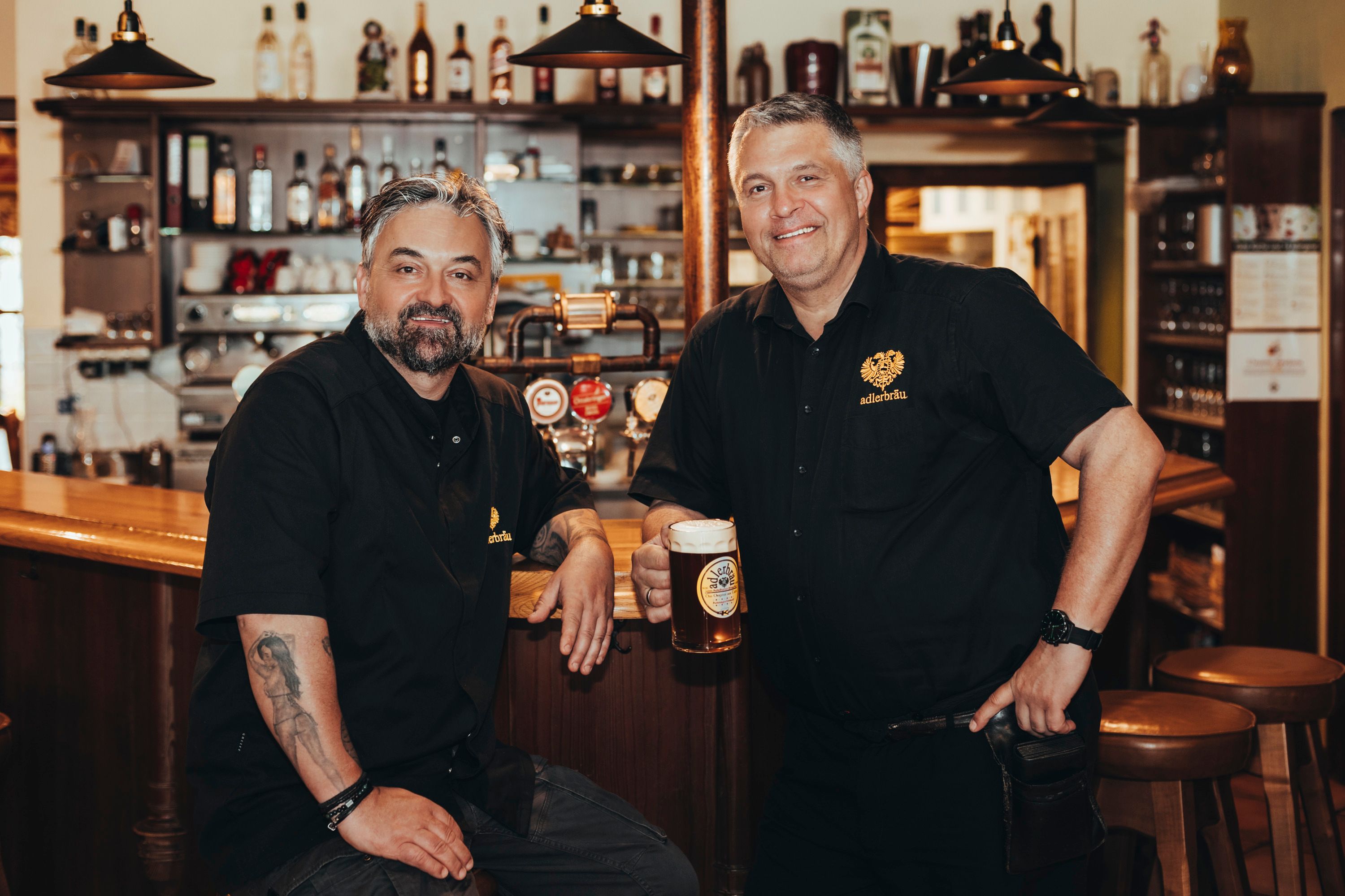 Two men dressed in black are standing in a bar with a beer mug in their hands.
