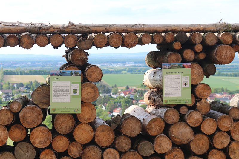 View through stacked logs onto a landscape with fields and houses.