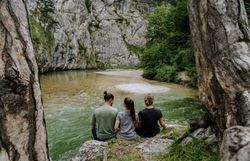Drei Personen sitzen am Ufer der Scharzach im Höllental am Wanderweg 1. Wiener Wasserleitungsweg und machen Pause. Rundum sie ragen Felden und Bäume in die Höhe. 